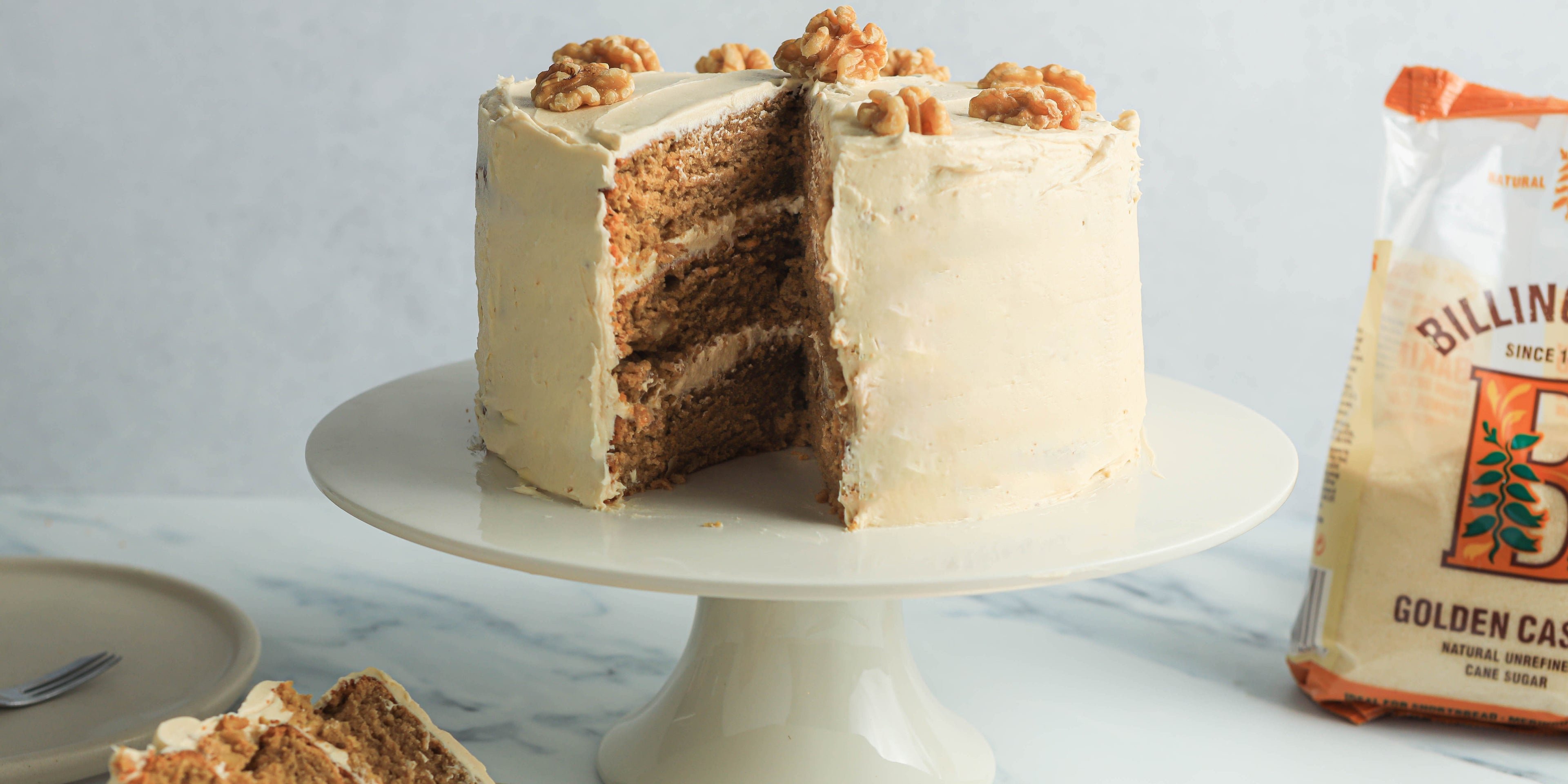Coffee and Walnut cake covered in buttercream and topped with walnuts on a white cake stand with a slice removed from it showing the layers inside. A pack of golden caster sugar next to it