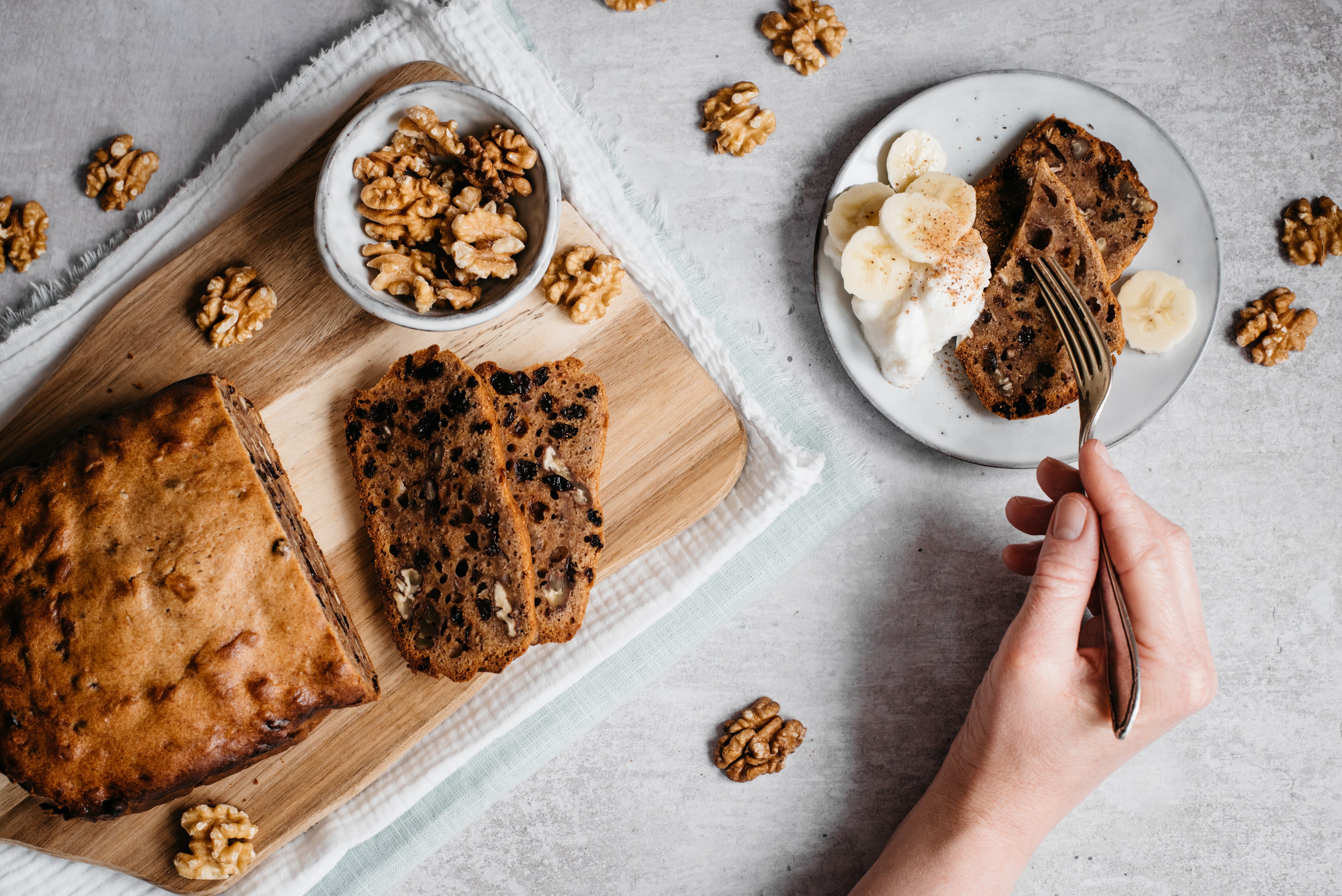 Overhead shot of banana bread loaf, slices infront, bowl of walnuts, plate with slices, fork and hand