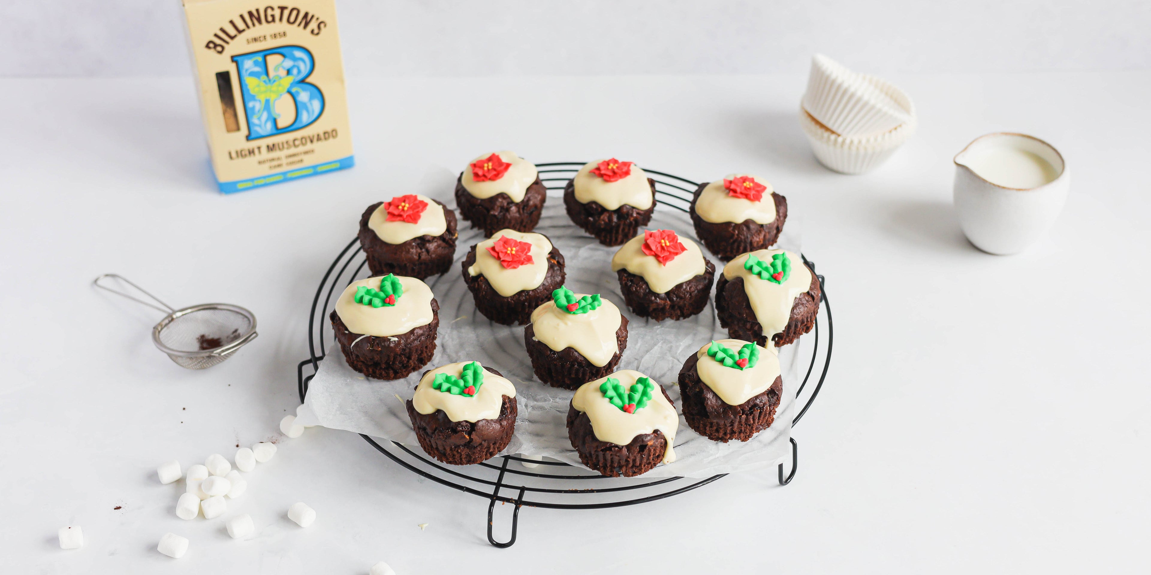 Top view of Mini Rocky Road Christmas Pudding Muffins on a wire rack, hand decorated with white chocolate. A pack of Billington's Light Muscovado sugar in the background