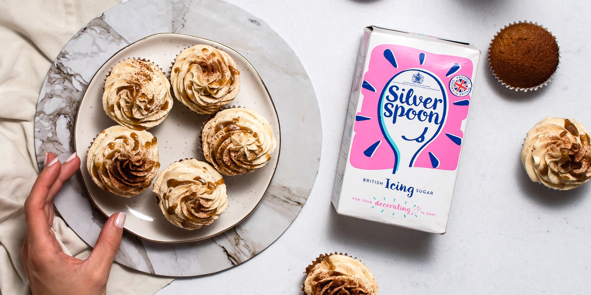 Top view of Gingerbread Cinnamon Cupcakes on a plate with a hand reaching for one. Next to a pack of Silver Spoon icing sugar