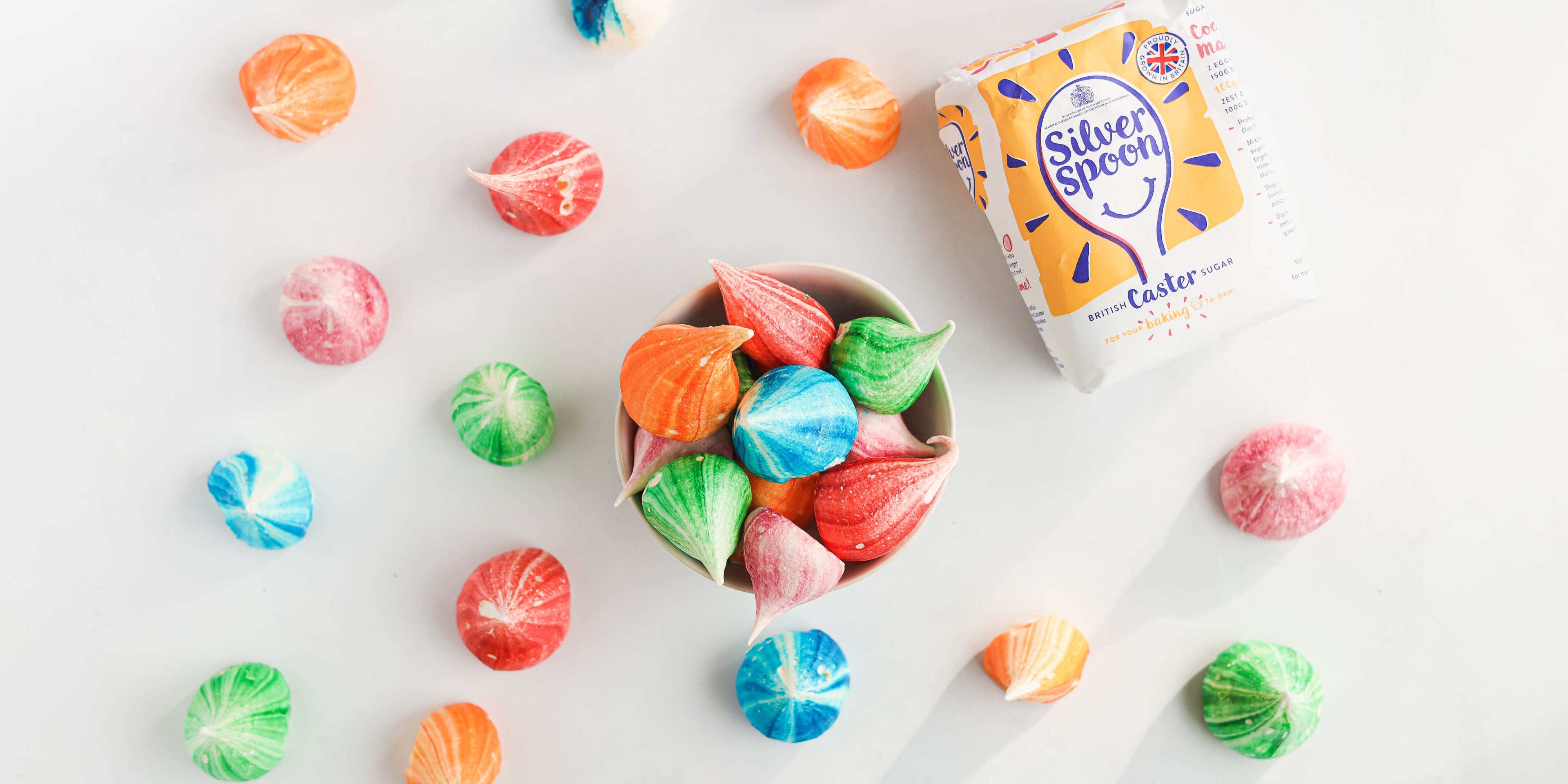 Top view of Rainbow Meringues in a bowl, with Rainbow Meringues scattered around next to a bag of silver spoon caster sugar