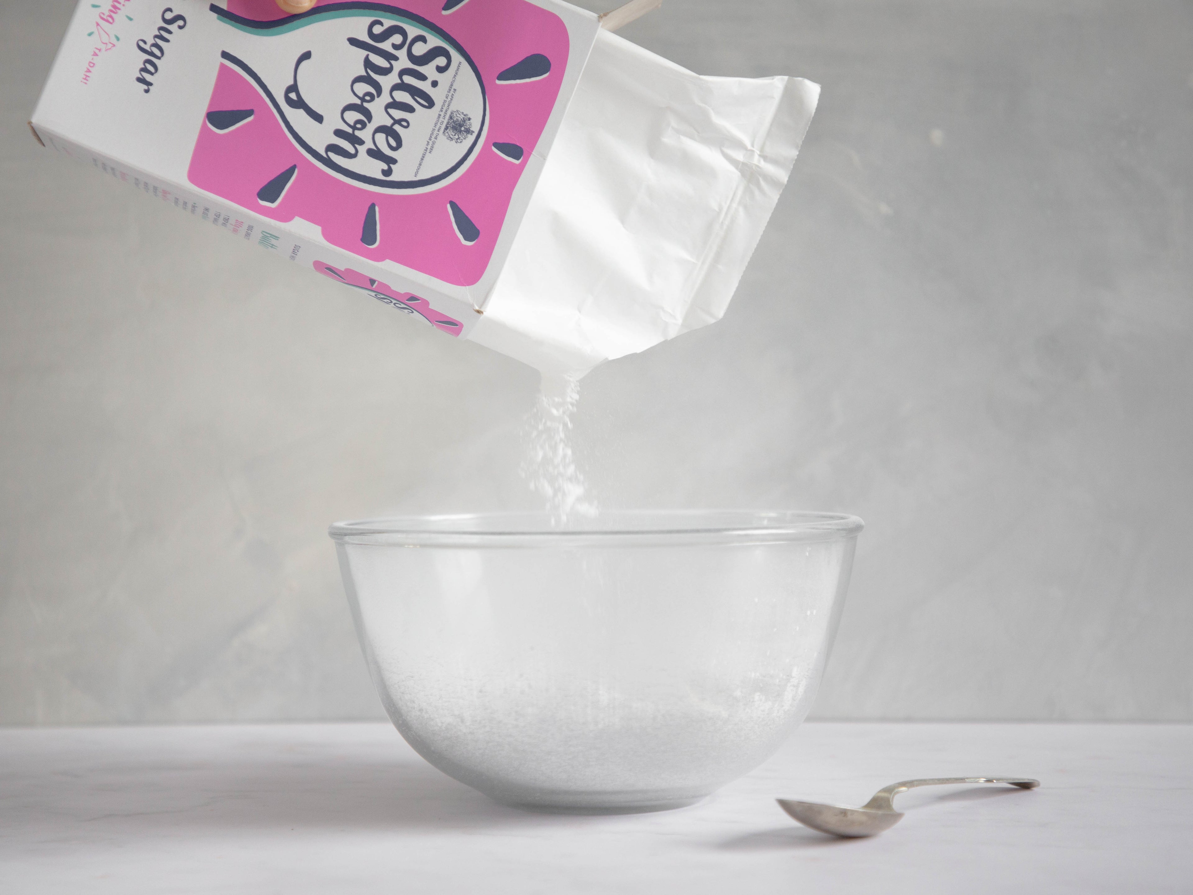 Box of icing sugar tipping sugar out into a glass bowl with a spoon on the side