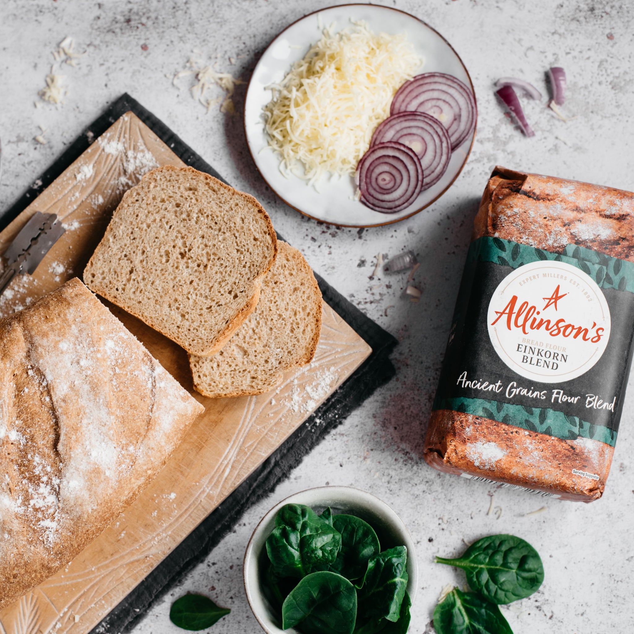 Overhead shot of einkorn bread with two slices in front. Plate with grated cheese, onion slices and spinach. Bread flour pack