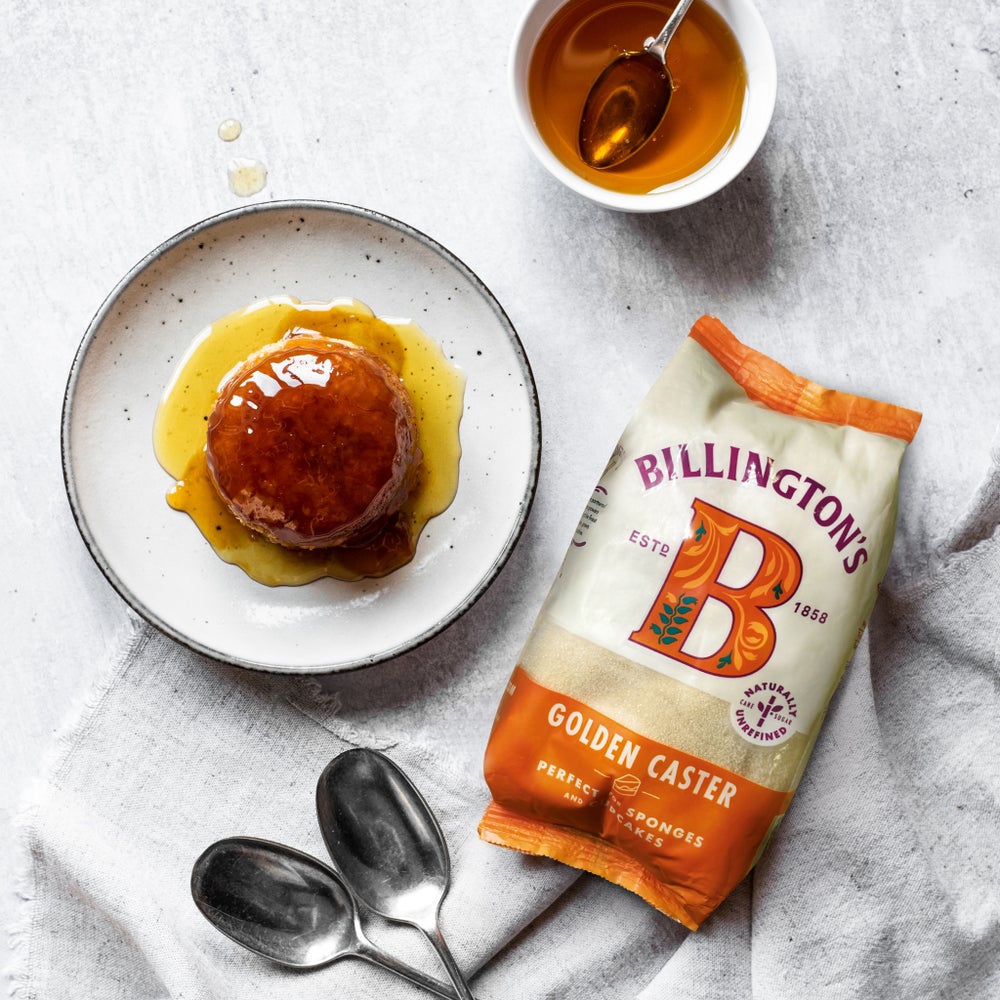 A bag of Billington's Golden Caster Sugar next to a microwaveable treacle sponge that's covered in syrup shot from above with a couple of spoons at the side and laid on a grey piece of linen.