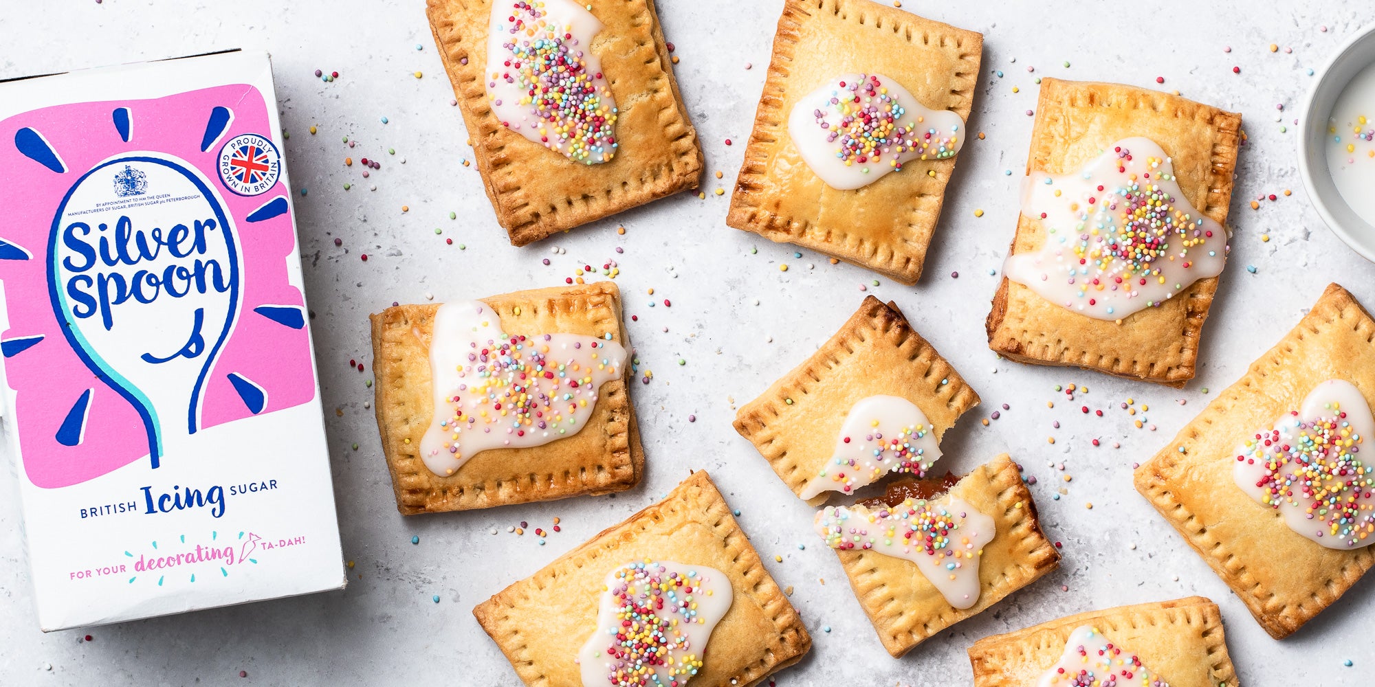 Overhead shot of Pop Tarts with white icing and sprinkles. One broken in half. Icing sugar pack in shot