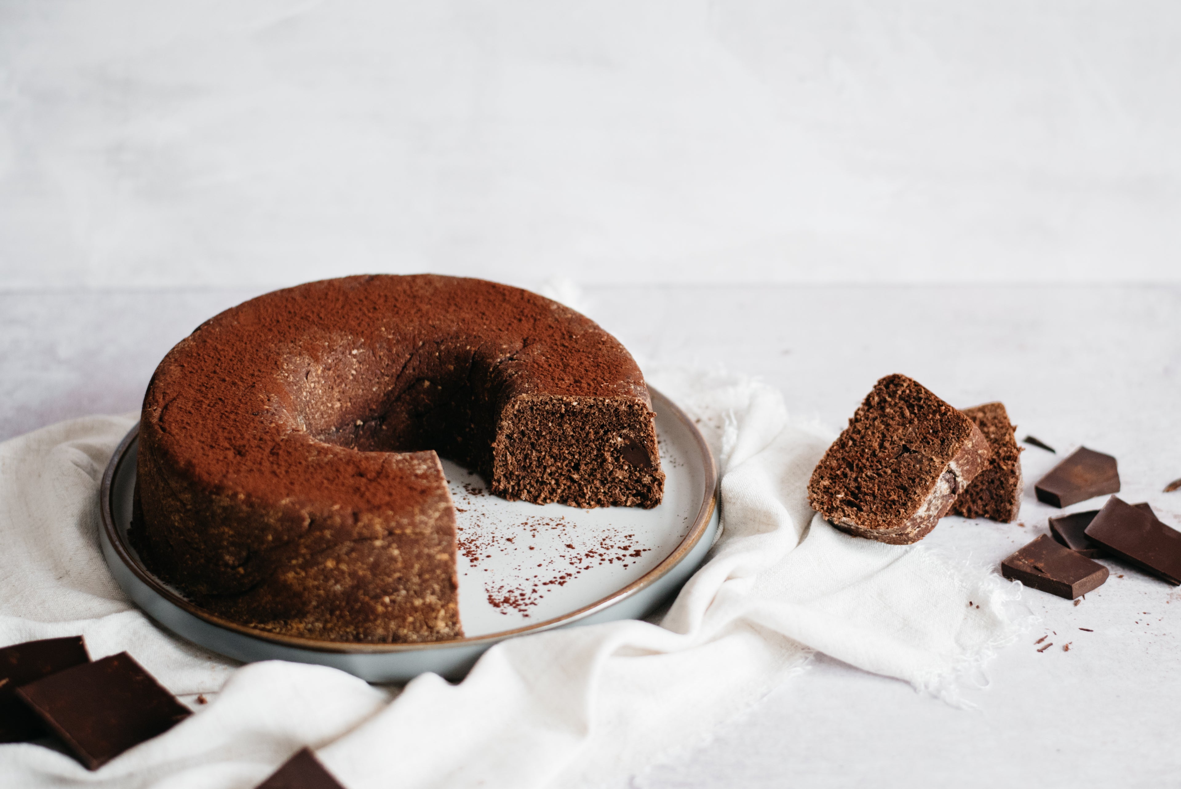 Chocolate Bread served on a plate, on a white cloth. Next to slices of Chocolate Bread stacked with pieces of chocolate