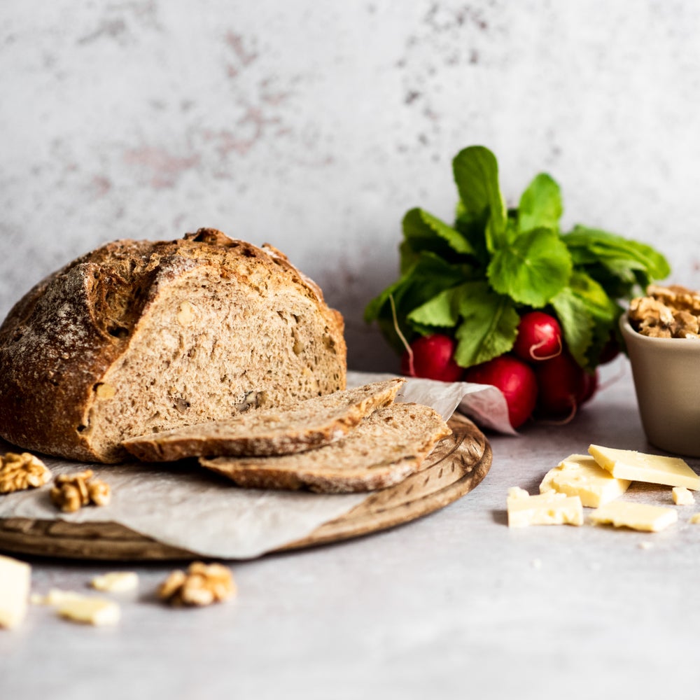 A loaf of chunky walnut bread with 2 slices cut and laid flat on a round wooden board. Some walnuts and slices of cheese are scattered in front of and next to the loaf.