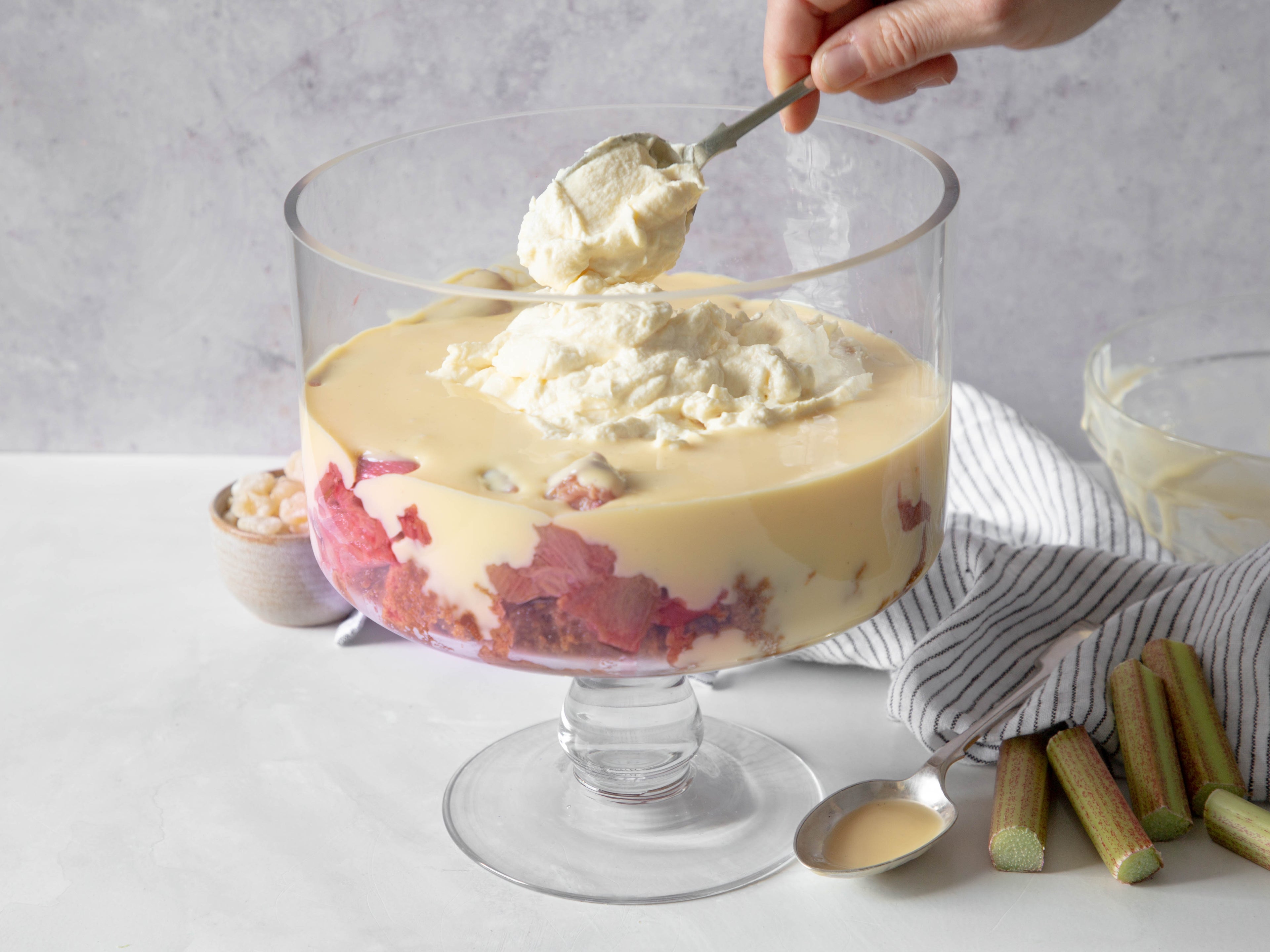 Trifle bowl filled with rhubarb and custard and a hand spooning in some cream on top. In the background there is a spoon, striped tea towel chopped rhubarb and a small bowl of nuts.