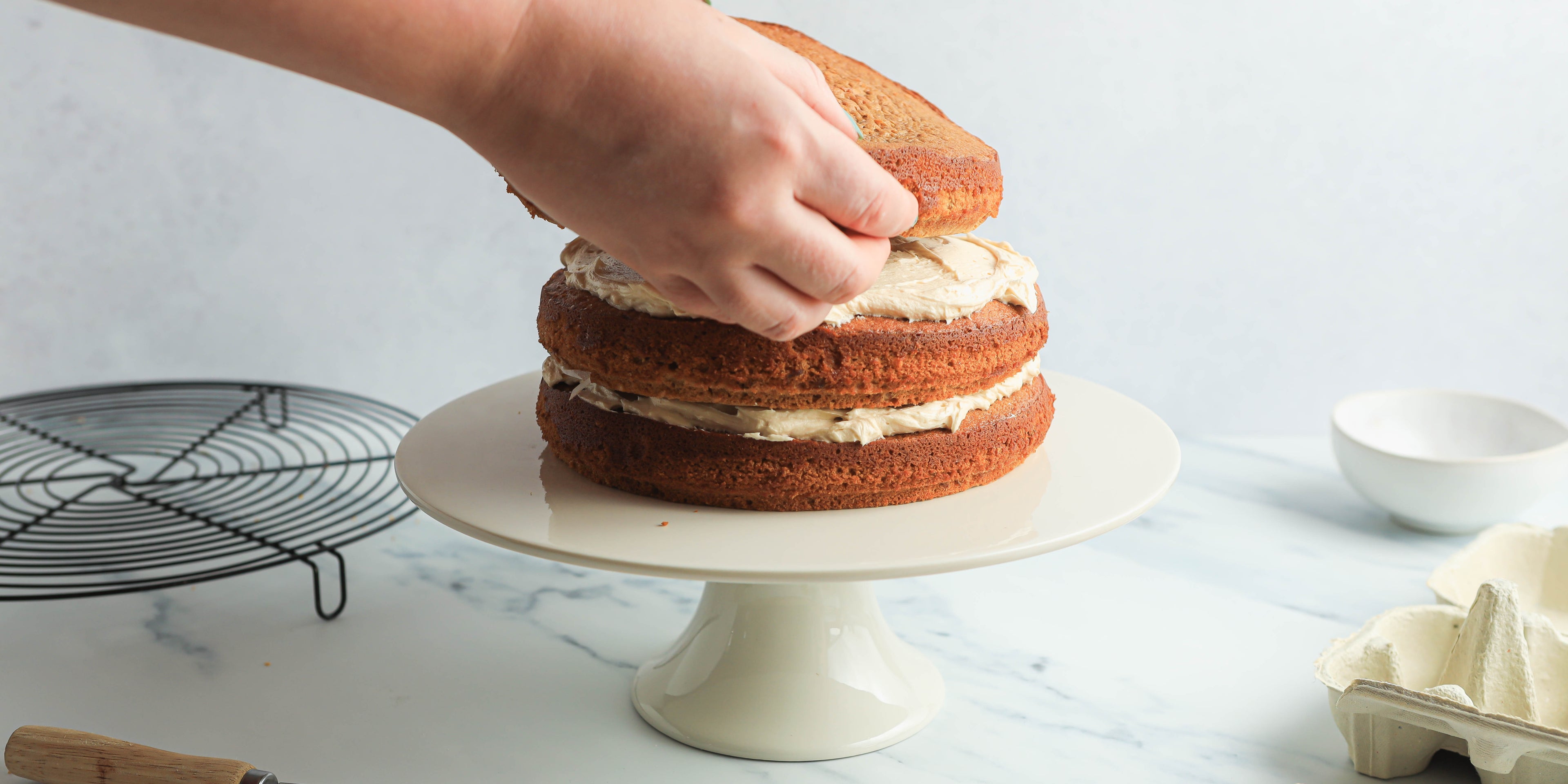 Two hands placing a layer of sponge on top of 2 sponge cakes with buttercream filling. The cake is on a white cake stand with a cooling rack next to it