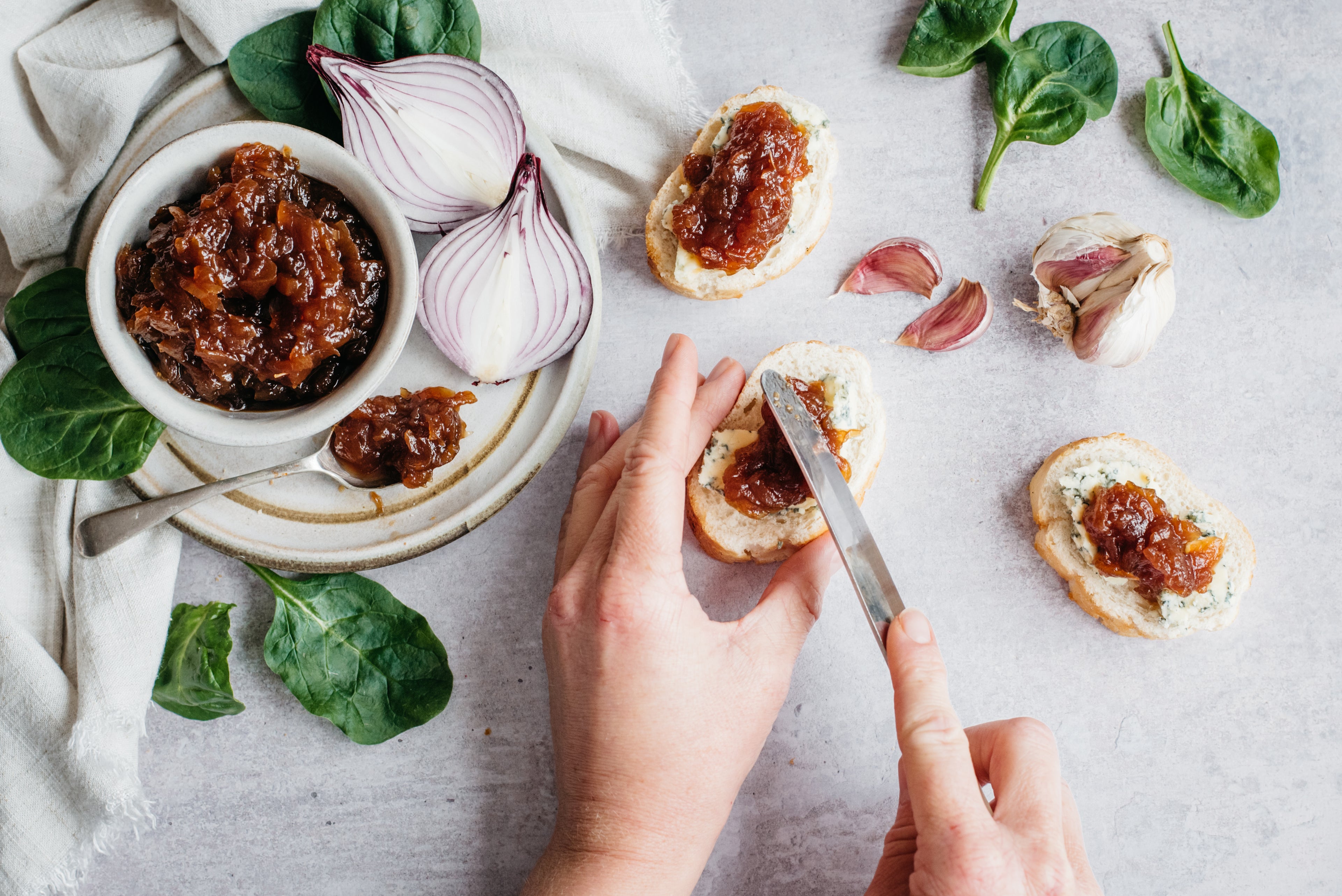 Overhead shot of hands spreading onion chutney on bread, herbs scattered around and pot of onion chutney with spoon