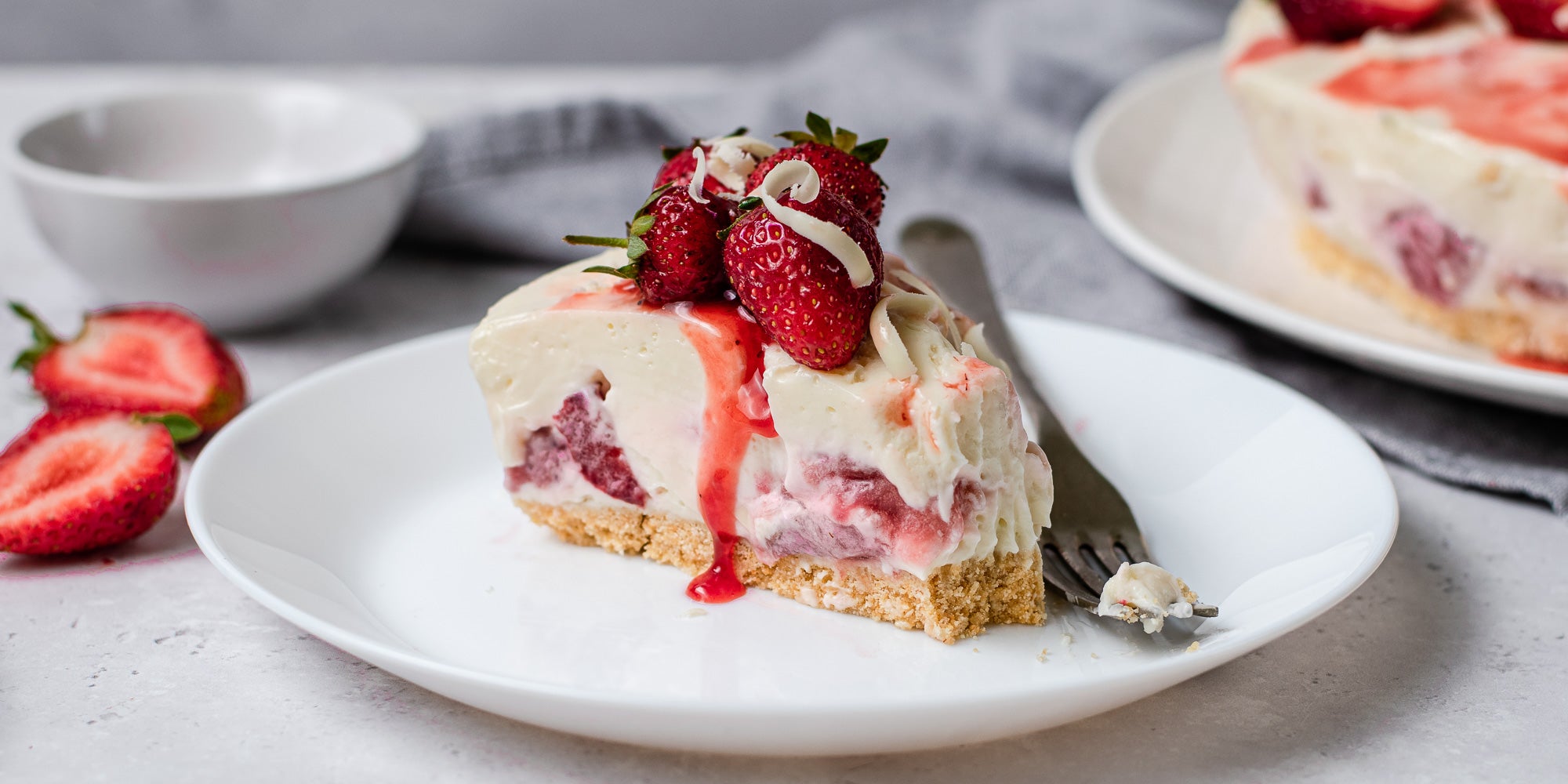 White Chocolate & Strawberry Cheesecake slice, with strawberry sauce running down the cheesecake. Next to a fork on a plate to serve.