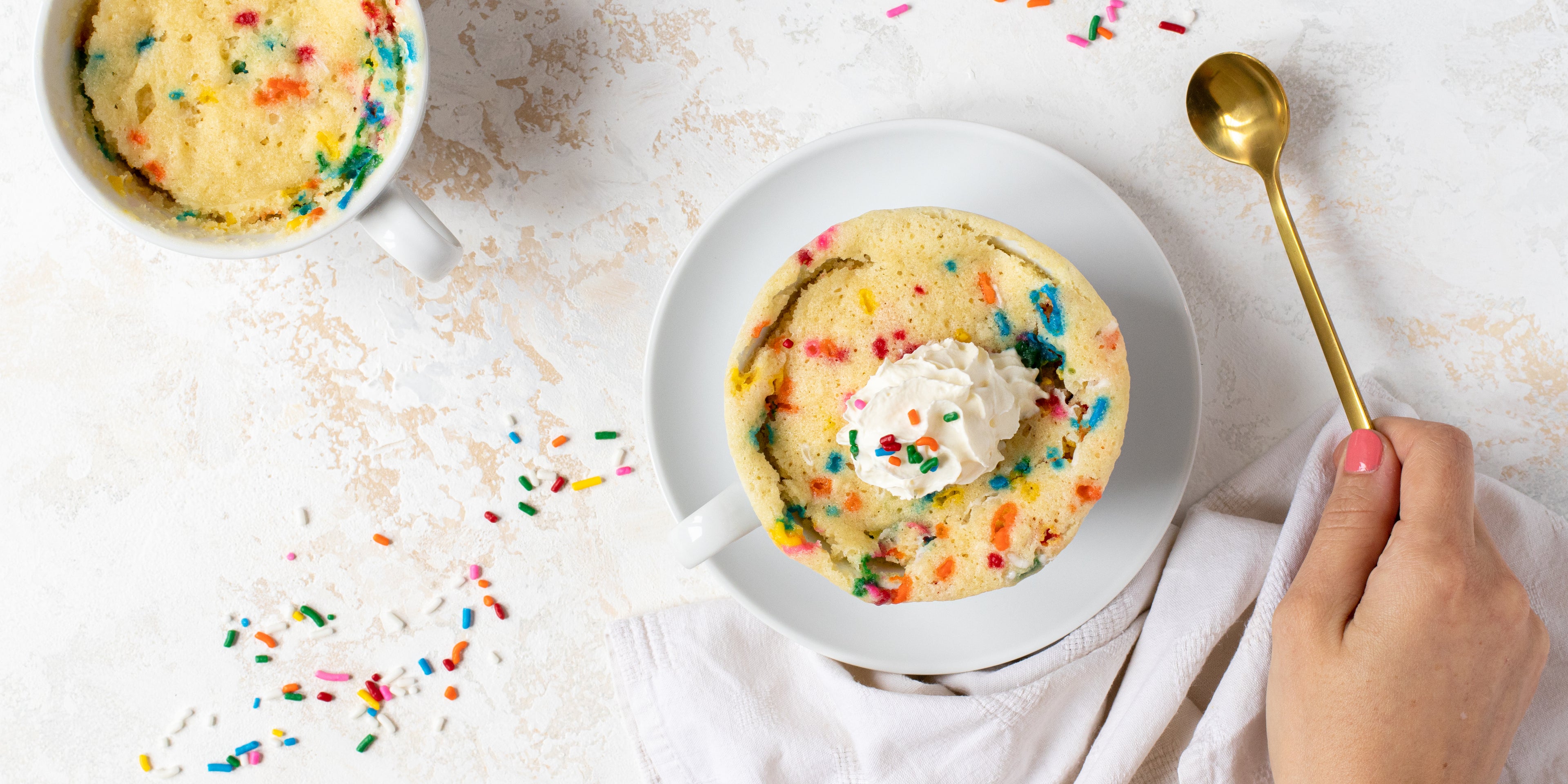 Birthday Cake Mug Cake with a hand holding a gold spoon. Mug cake is topped with whipped cream and sprinkles