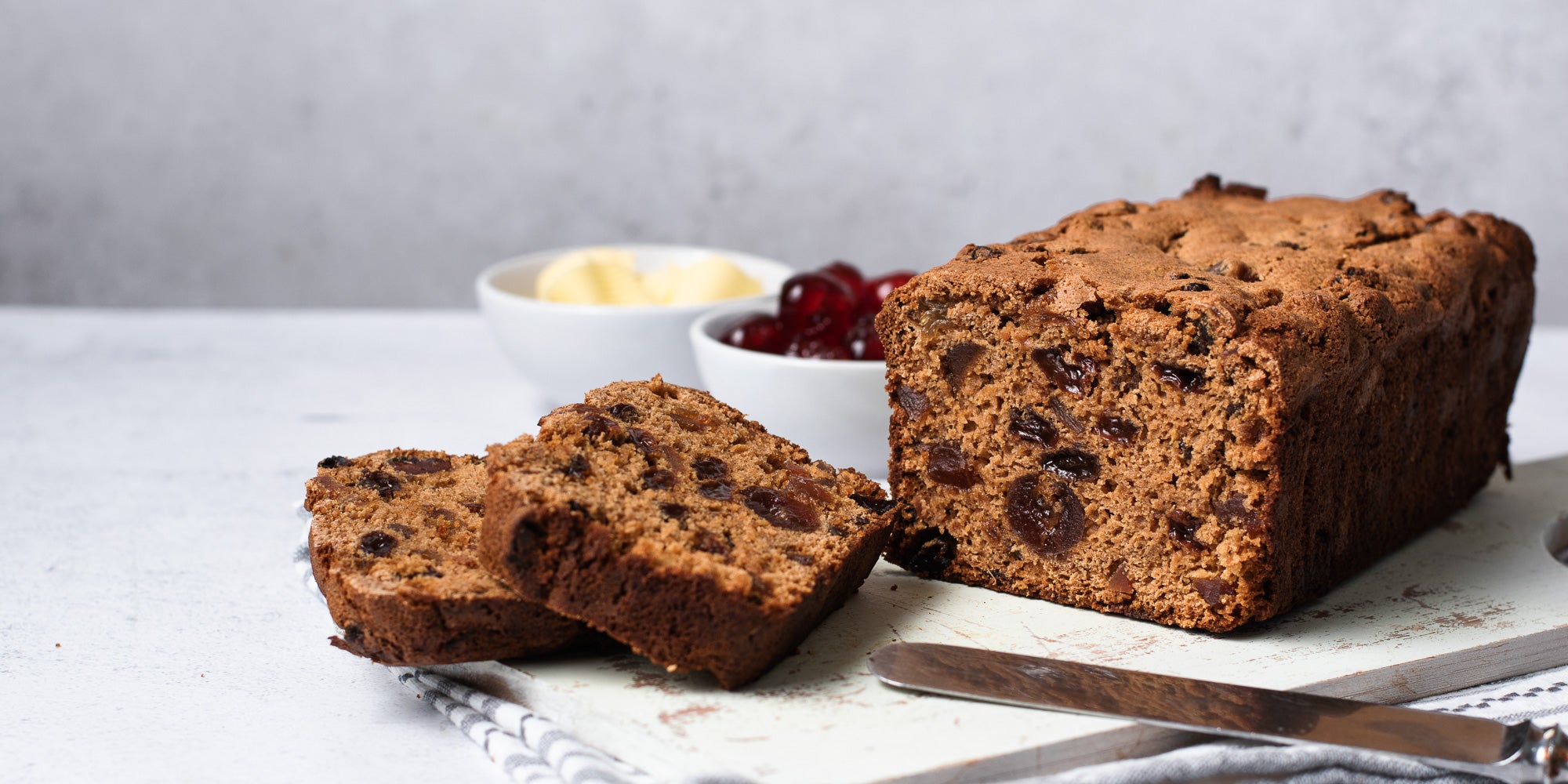 Irish tea loaf with two slices infront and pots of butter and cherries. Knife in shot