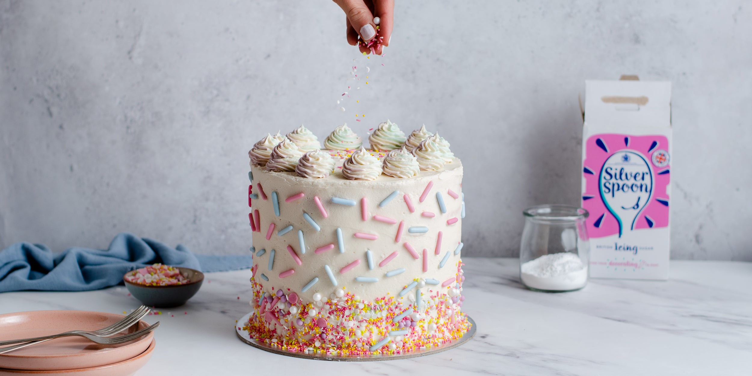 Sponge cake coated in buttercream and rainbow sprinkles. Hand above adding sprinkles. Icing sugar pack to the side. Plates and crockery to the left