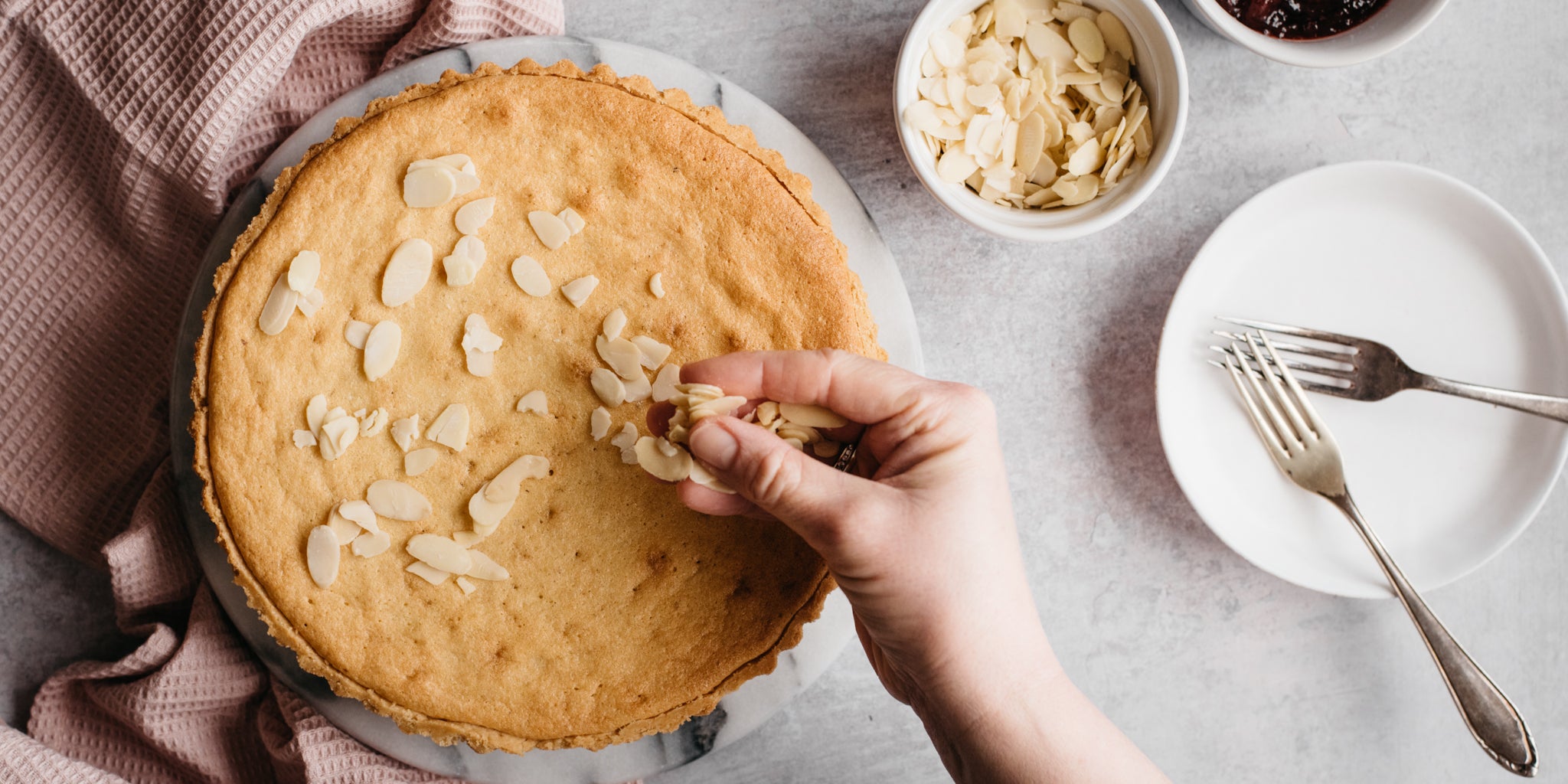 Hand sprinkling Simple Bakewell Tart with toasted almonds, next to a dish of almonds and forks to serve