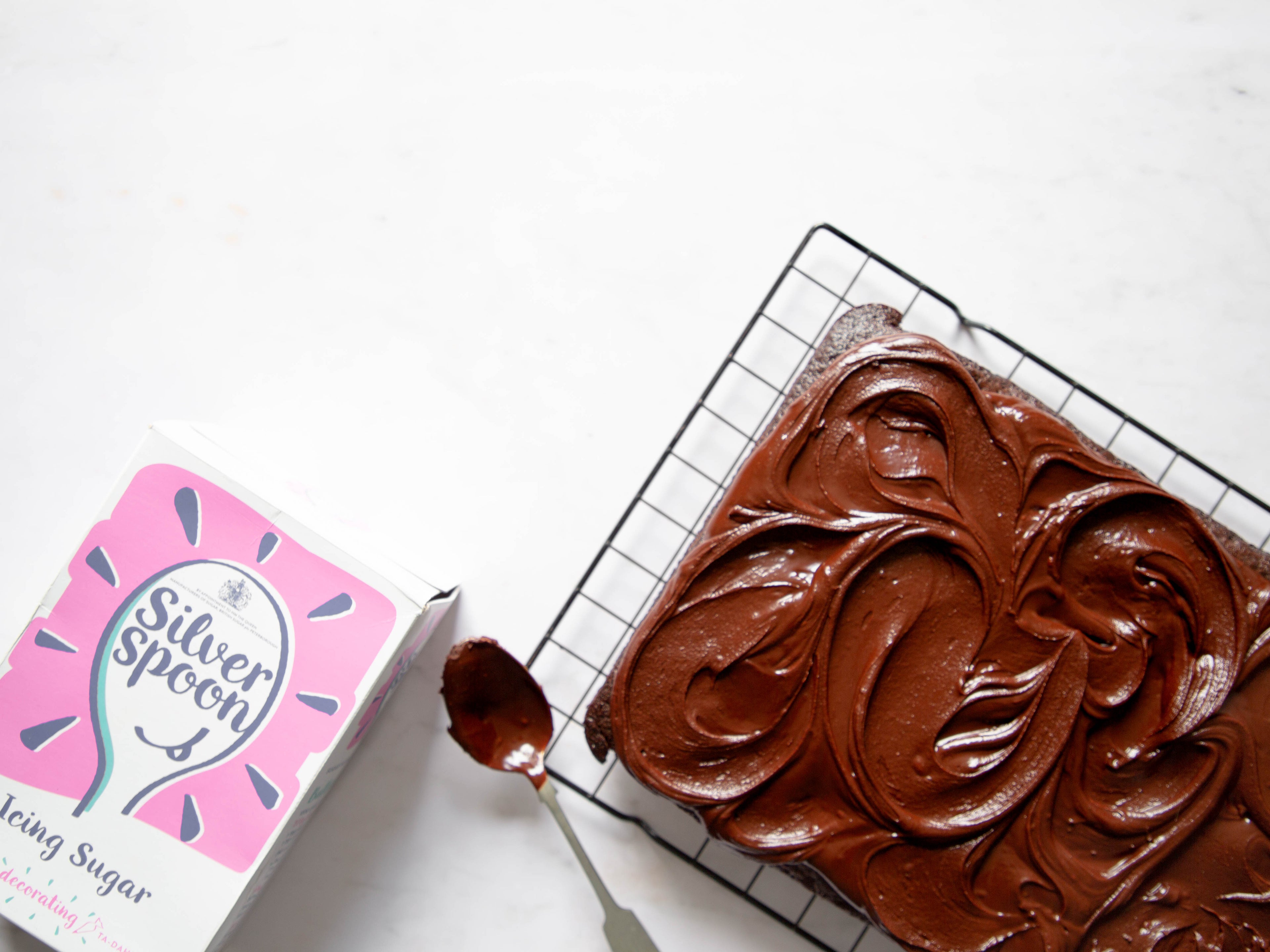 Box of icing sugar on the left, next to a chocolate covered sponge and chocolate traybake topped with chocolate icing on a cooling rack.  