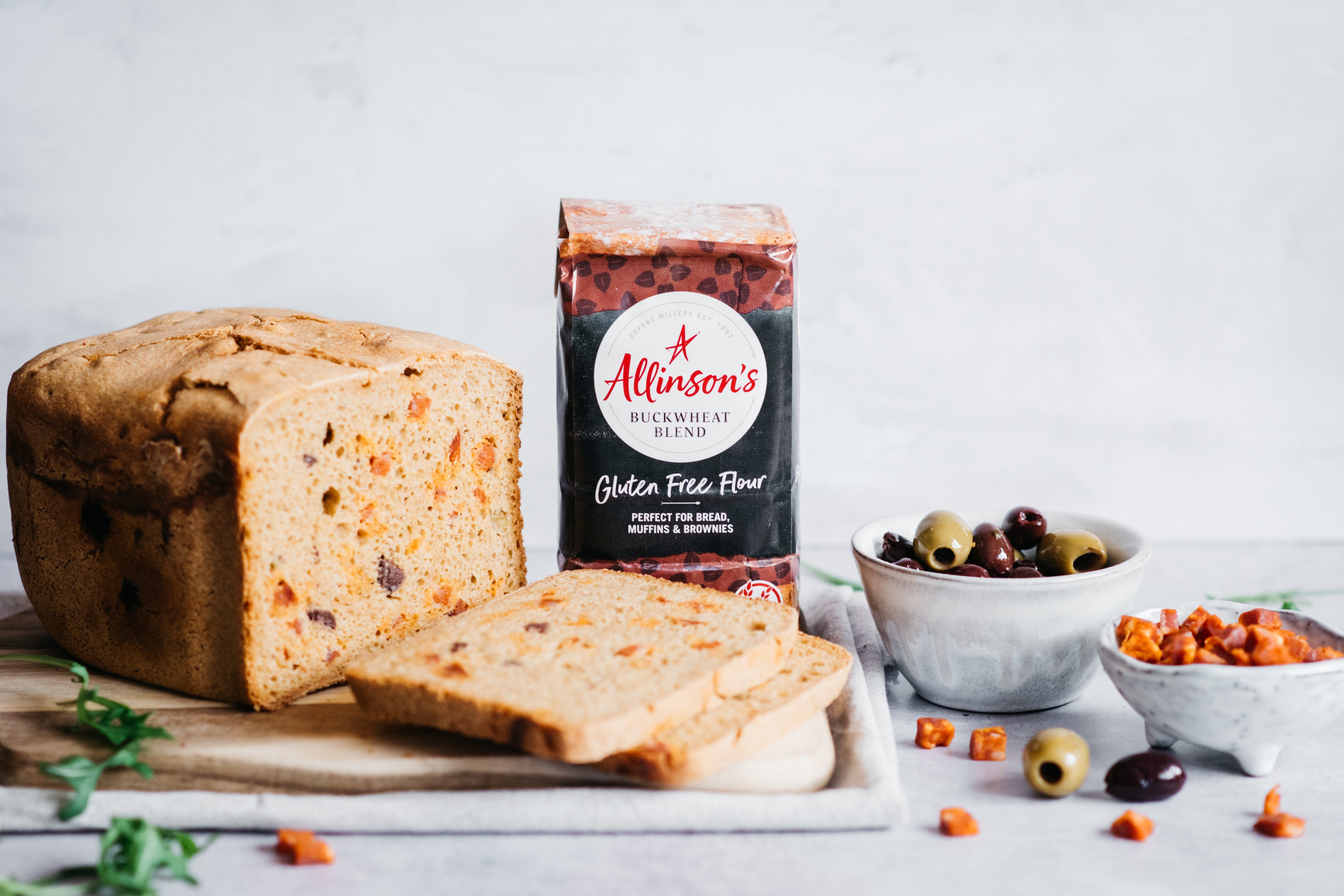 Loaf of bread on chopping board with slices in front, bread flour pack and two bowls of olives and chorizo