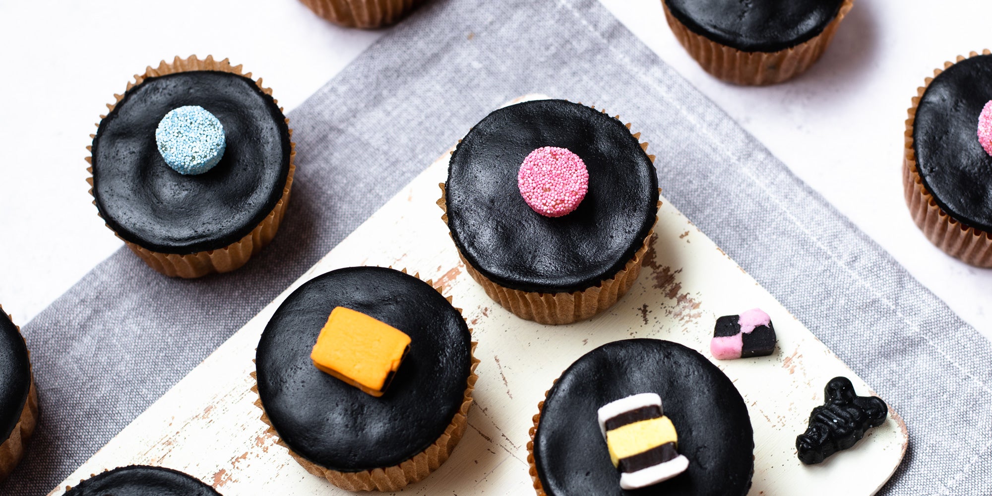 Close up of four cupcakes in brown cases, black fondant, topped with liquorice allsorts. Sweets dotted around