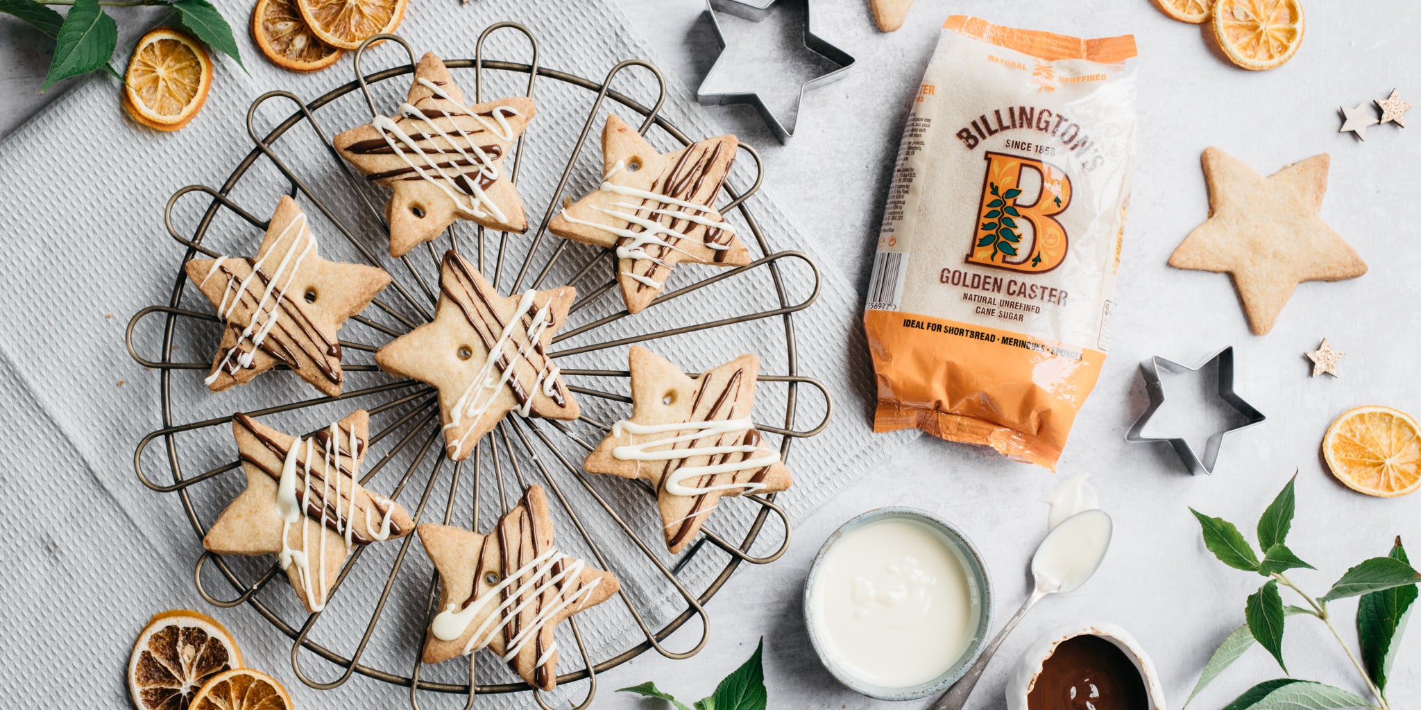 Top view of Orange & Ginger shortbread stars next to a bag of Billington's golden caster sugar, cookie cutters and melted chocolate on a spoon