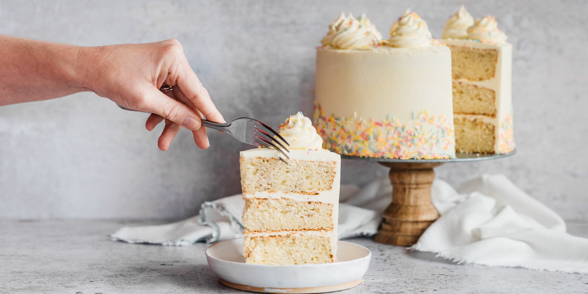 Vegan Vanilla Cake with a slice cut out of it, served on a plate with a fork cutting in to take a slice. 