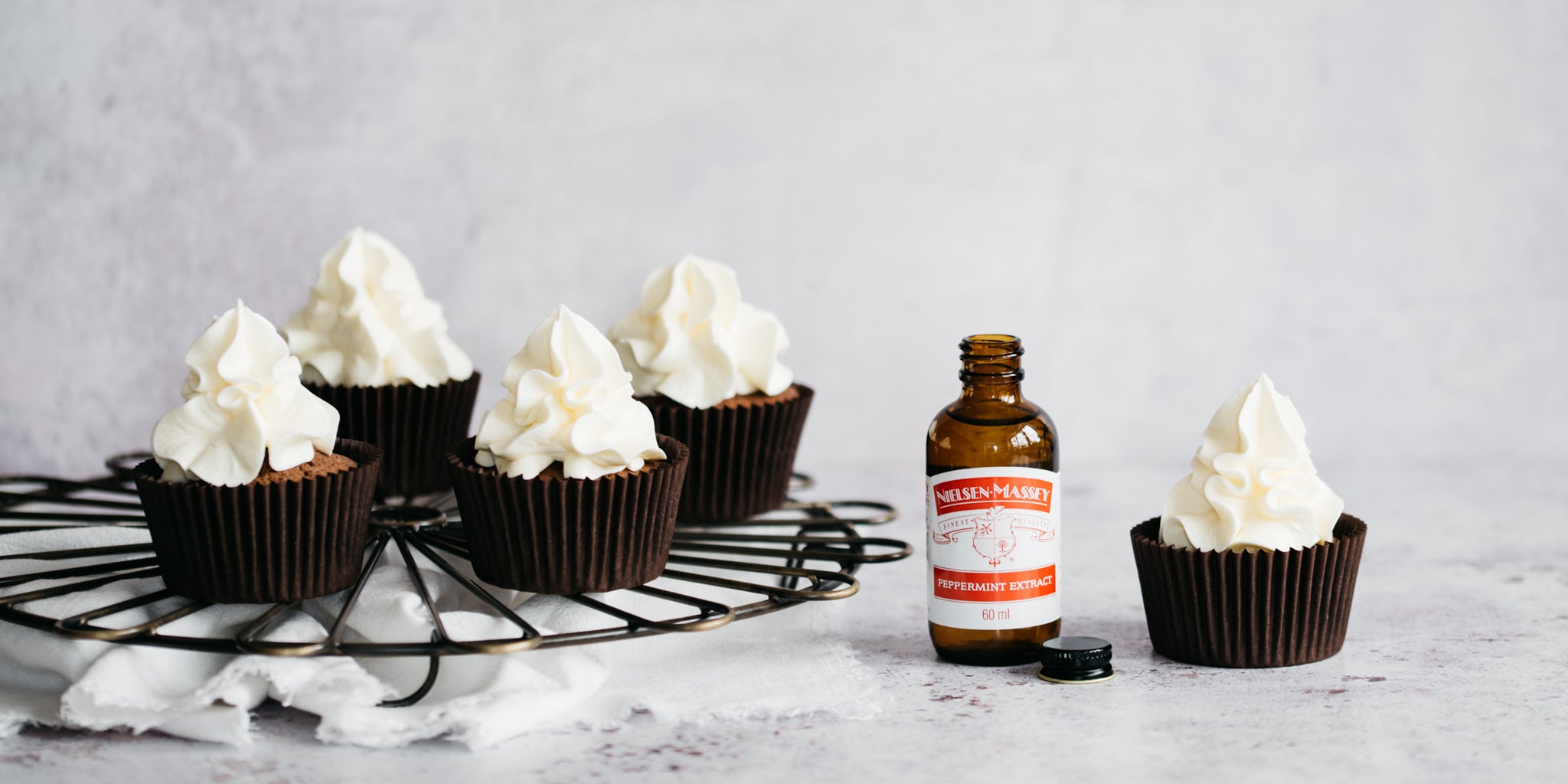 Chocolate & Peppermint Cupcakes on a wire rack, swirled with peppermint buttercream next to a bottle of Nielsen Massey Peppermint Extract