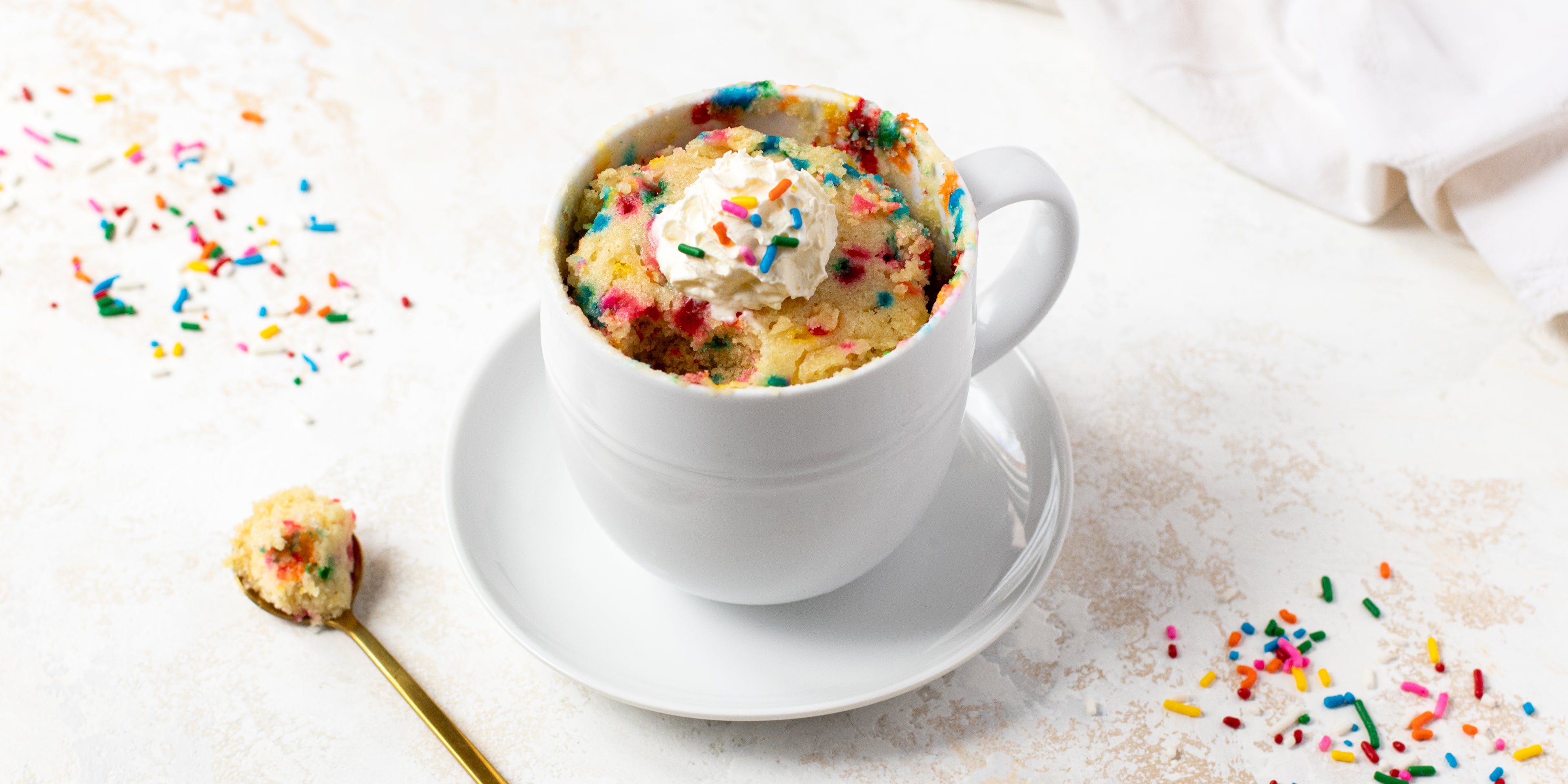 Birthday Cake Mug Cake with a spoon taken out of it, showing inside the mug cake. Surrounded by sprinkles and a spoon with cake on it