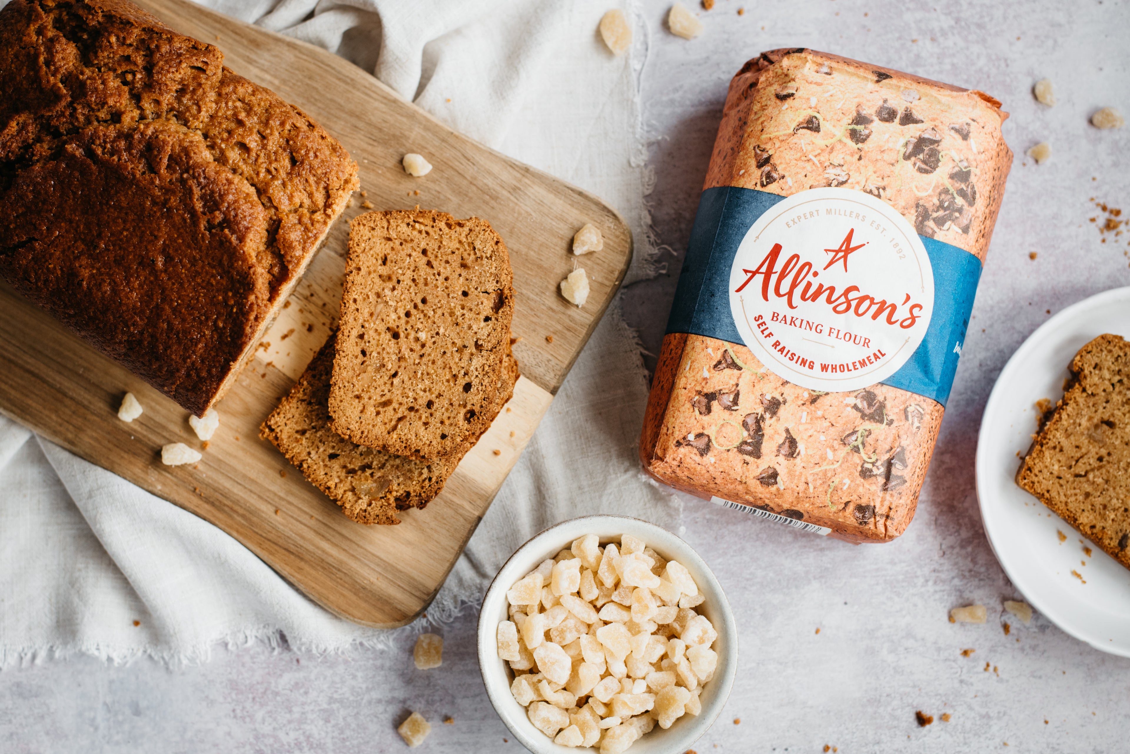 Top view of Gingerbread Loaf on a wooden serving board, next to a bowl of crystallised ginger and a bag of Allinson's self raising flour