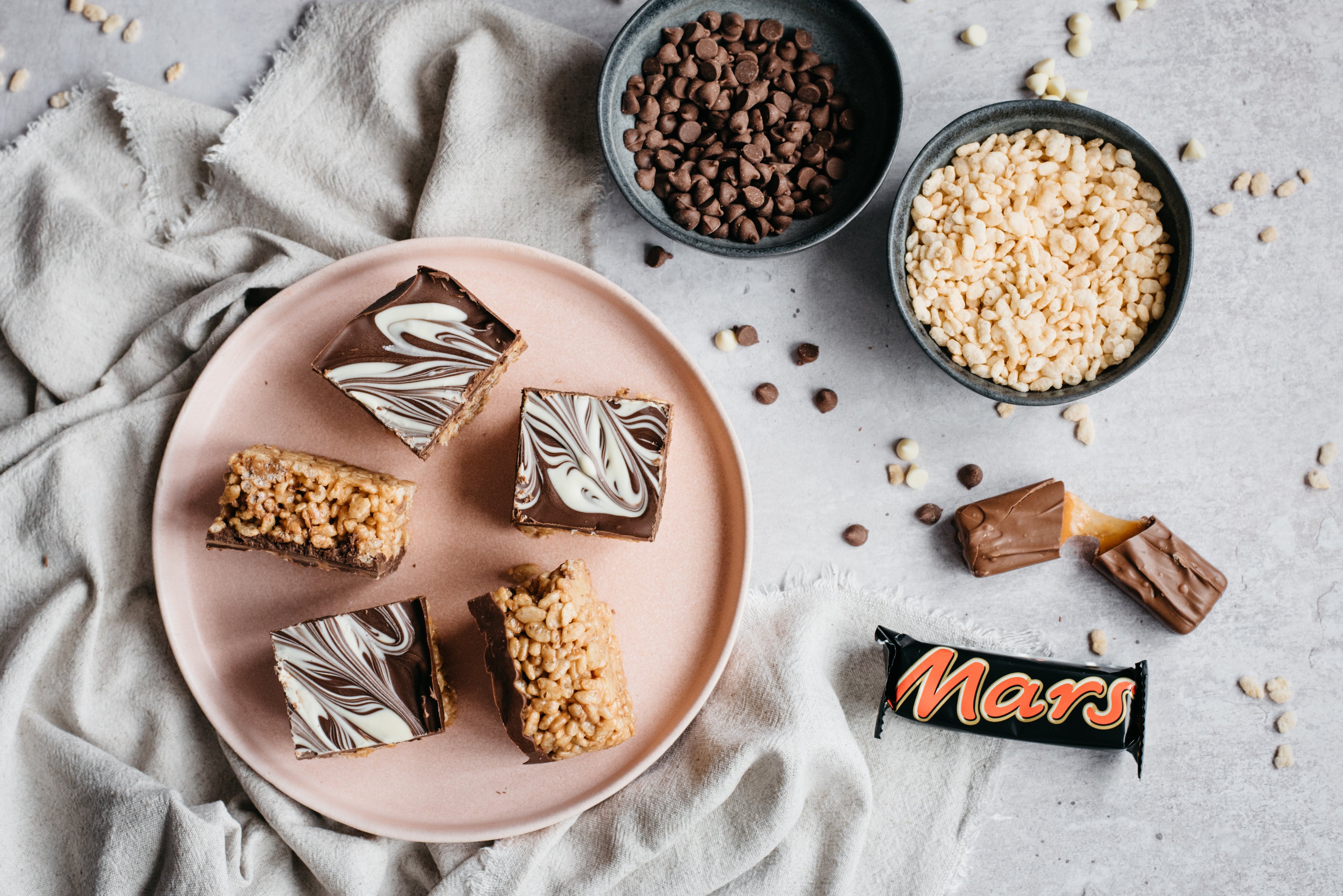 Homemade Mars Bar Cakes on a pink plate with a Mars Bar, bowl of rice krispies and chocolate chips
