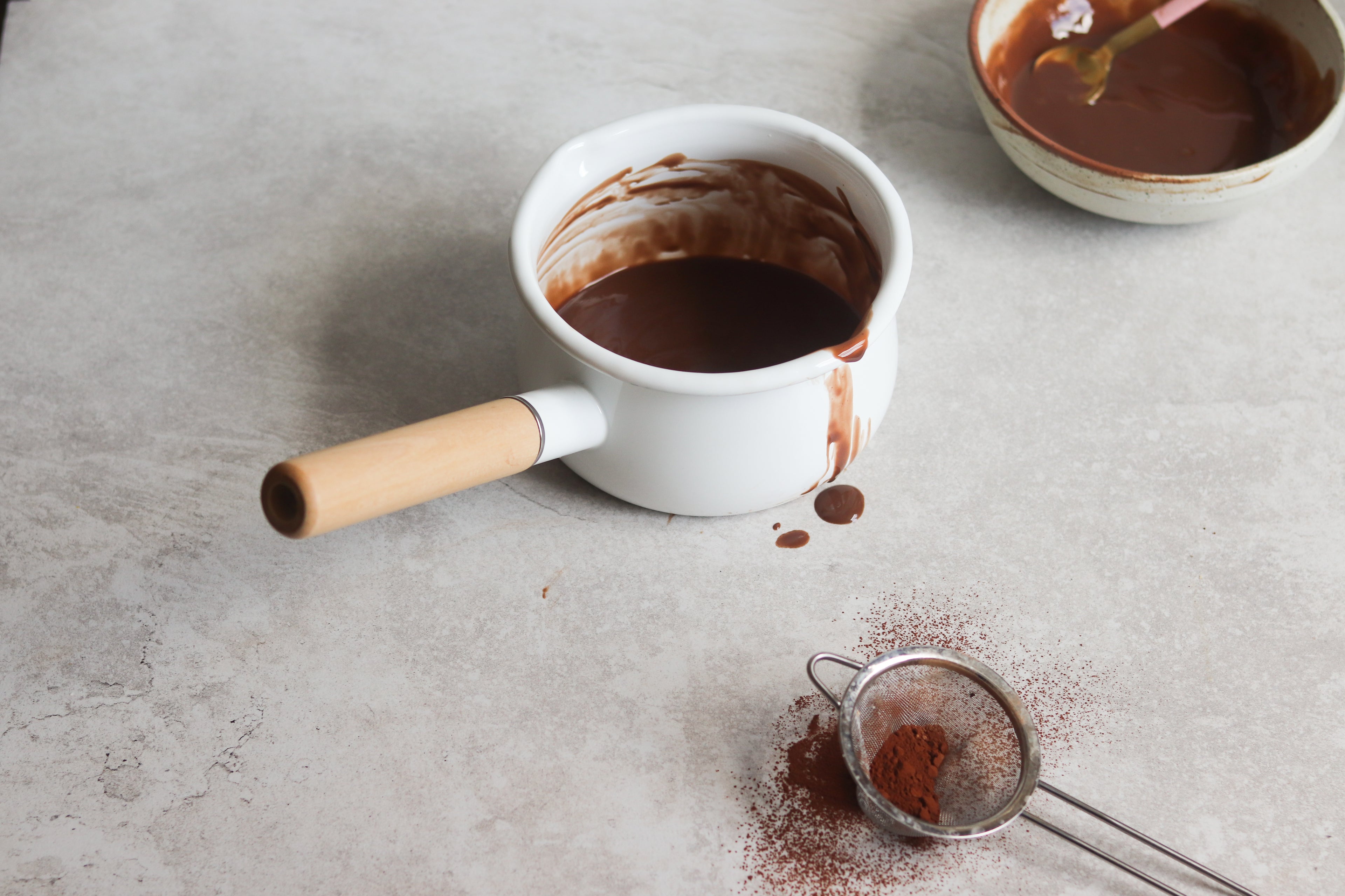 Saucepan of Chocolate Custard with drips down the side. Next to a sieve of cocoa powder