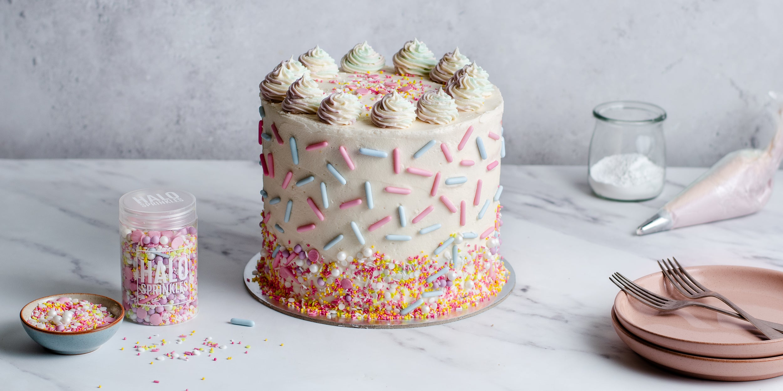 Cake covered in buttercream and sprinkles. Pot of sprinkles beside it along with piping bag and crockery