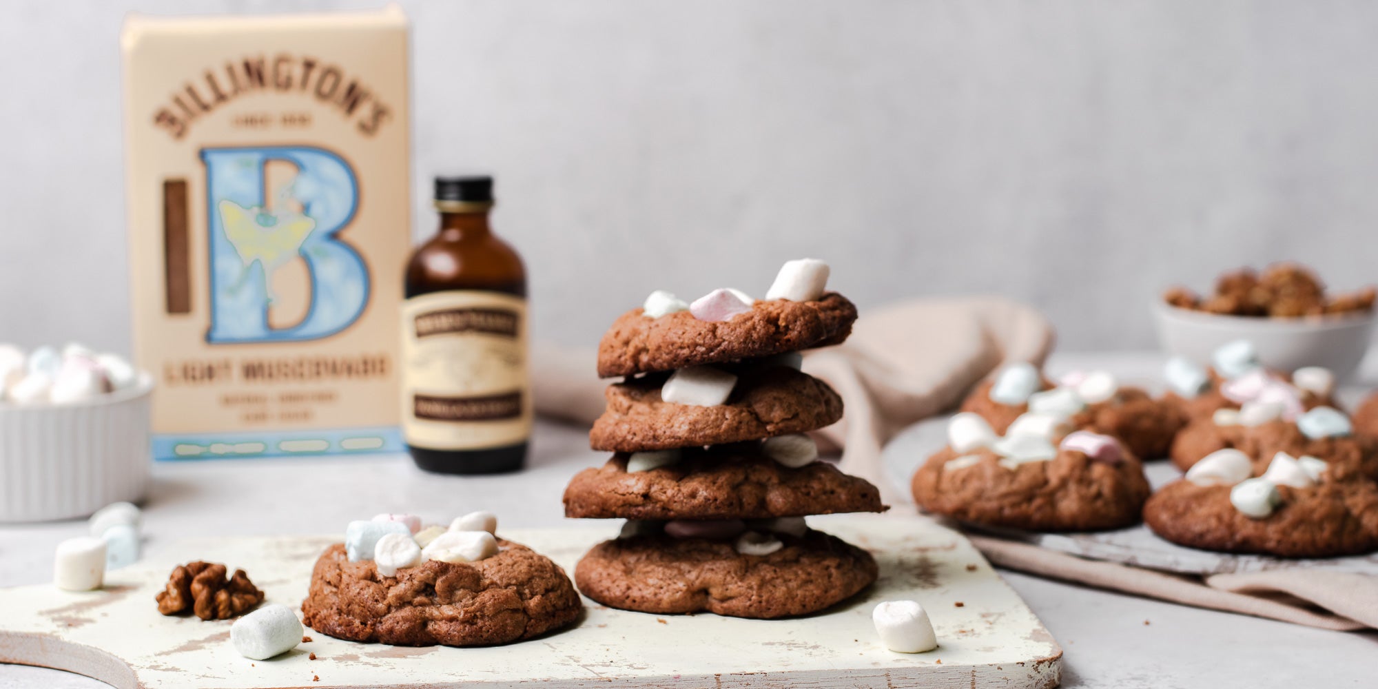 Rocky Road Cookies stacked on top of each other on a serving board, next to a bottle of Neilsen-Massey Vanilla extract, with a box of Billington's Light Muscovado sugar behind