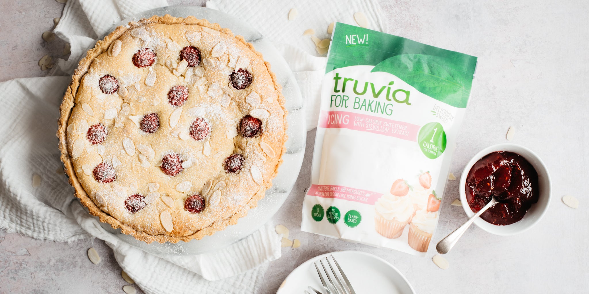 Bakewell tart on a white plate with a pack of Truvia for baking icing sugar and a pot of jam beside it