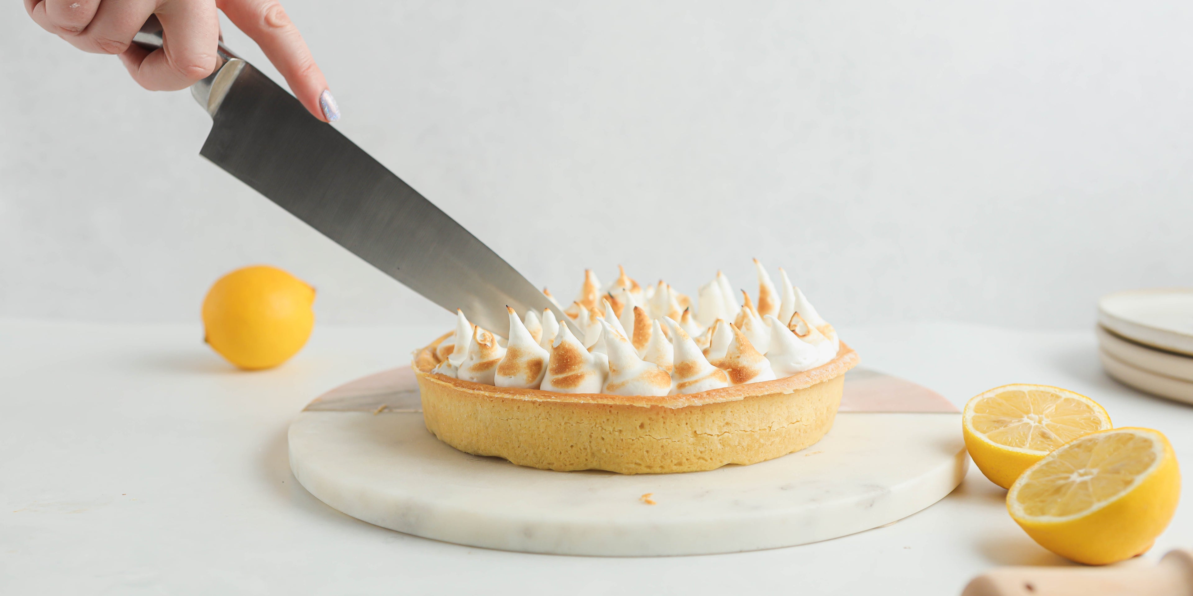 Lemon meringue pie on circular plate with knife cutting into it. Lemon slices at the side and a stack of white plates