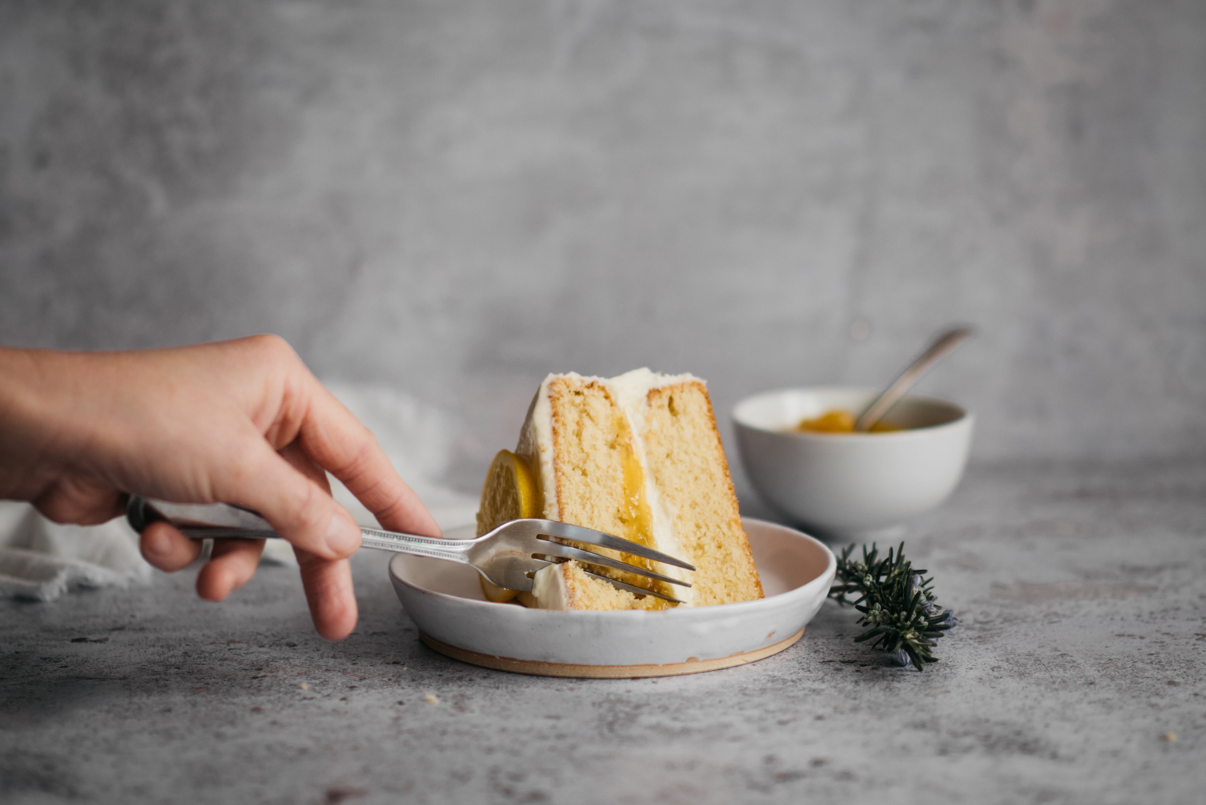 Hand cutting into a slice of Gluten Free Elderflower & Lemon Cake with a fork, showing the lemon and buttercream filling.