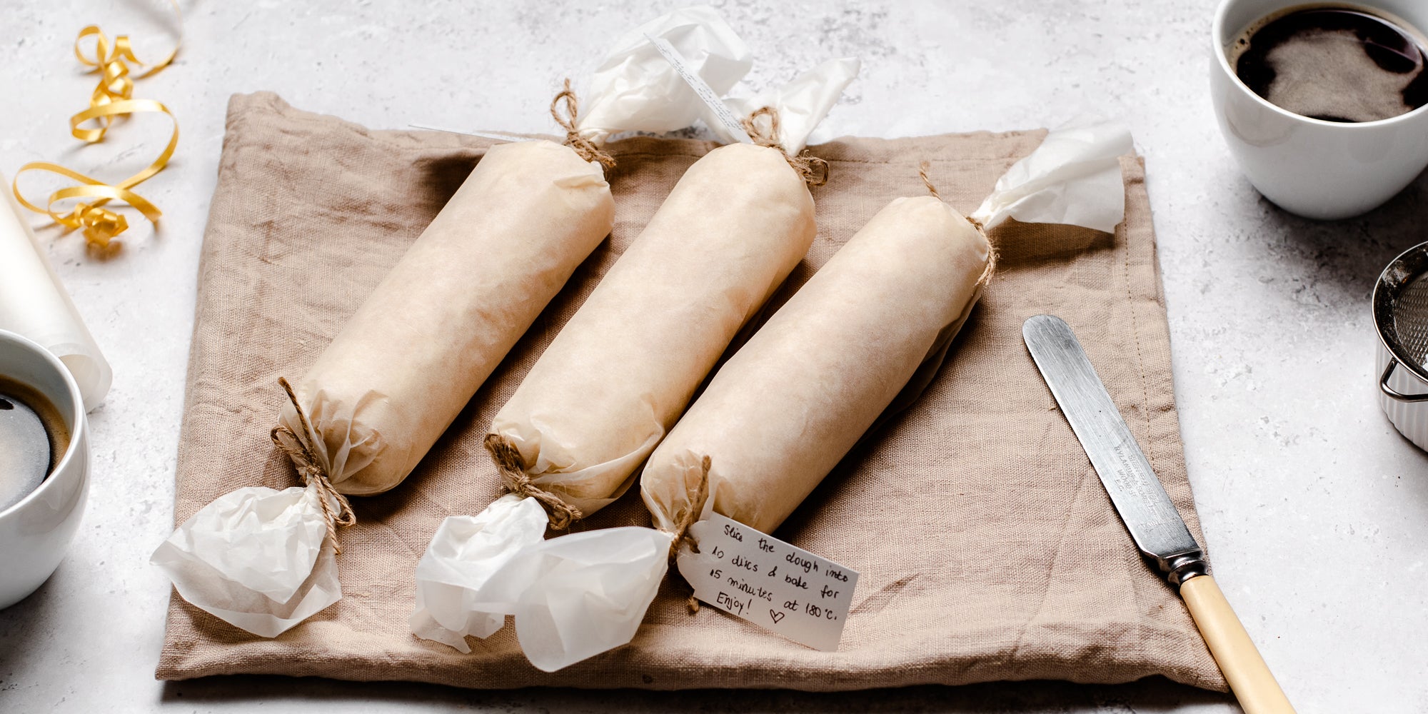 Shortbread Dough rolls wrapped in parchment and tied with a gold ribbon lay on a linen cloth. The rolls have handwritten notes to give the dough as a gift