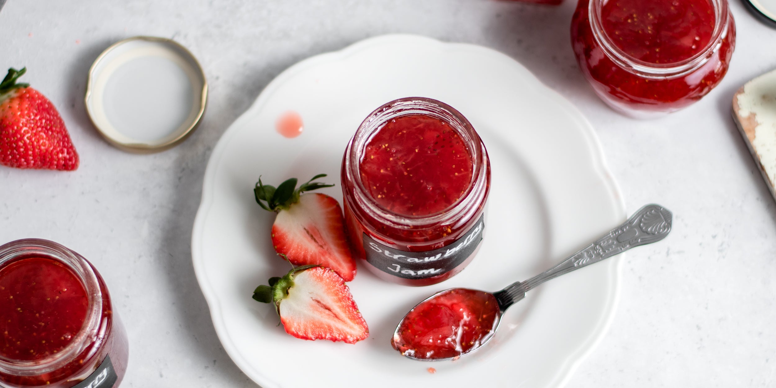 Top view of a jar of Strawberry Jam next to a spoon with a spoonful of jam and sliced strawberries