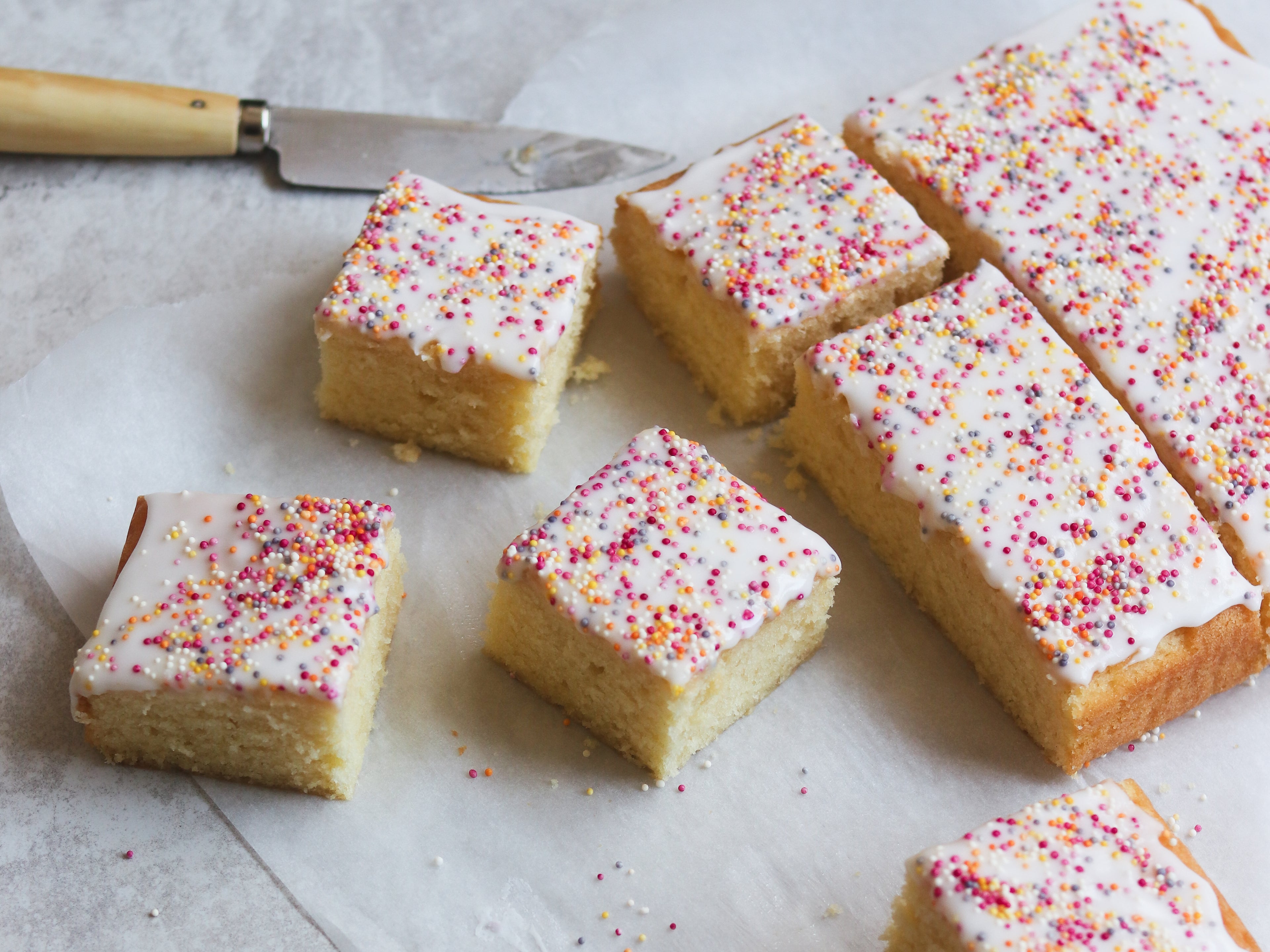 Sprinkle Cake sliced up on a sheet of baking paper, topped with icing and sprinkles