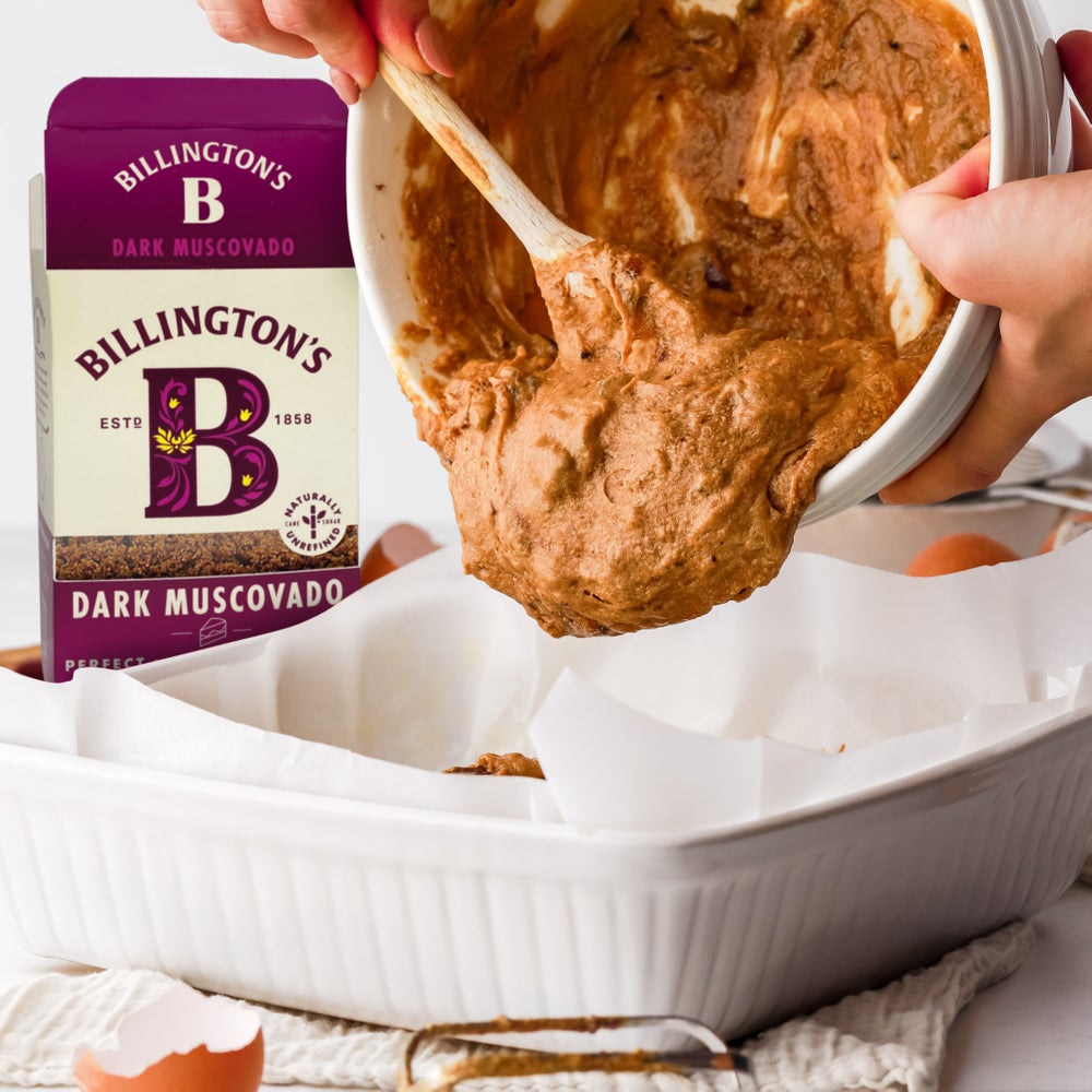 A box of Billington's Dark Muscovado Sugar next to a white ceramic dish which is about to have sticky toffee pudding batter spooned into it by someone using a wooden spoon and a large mixing bowl.