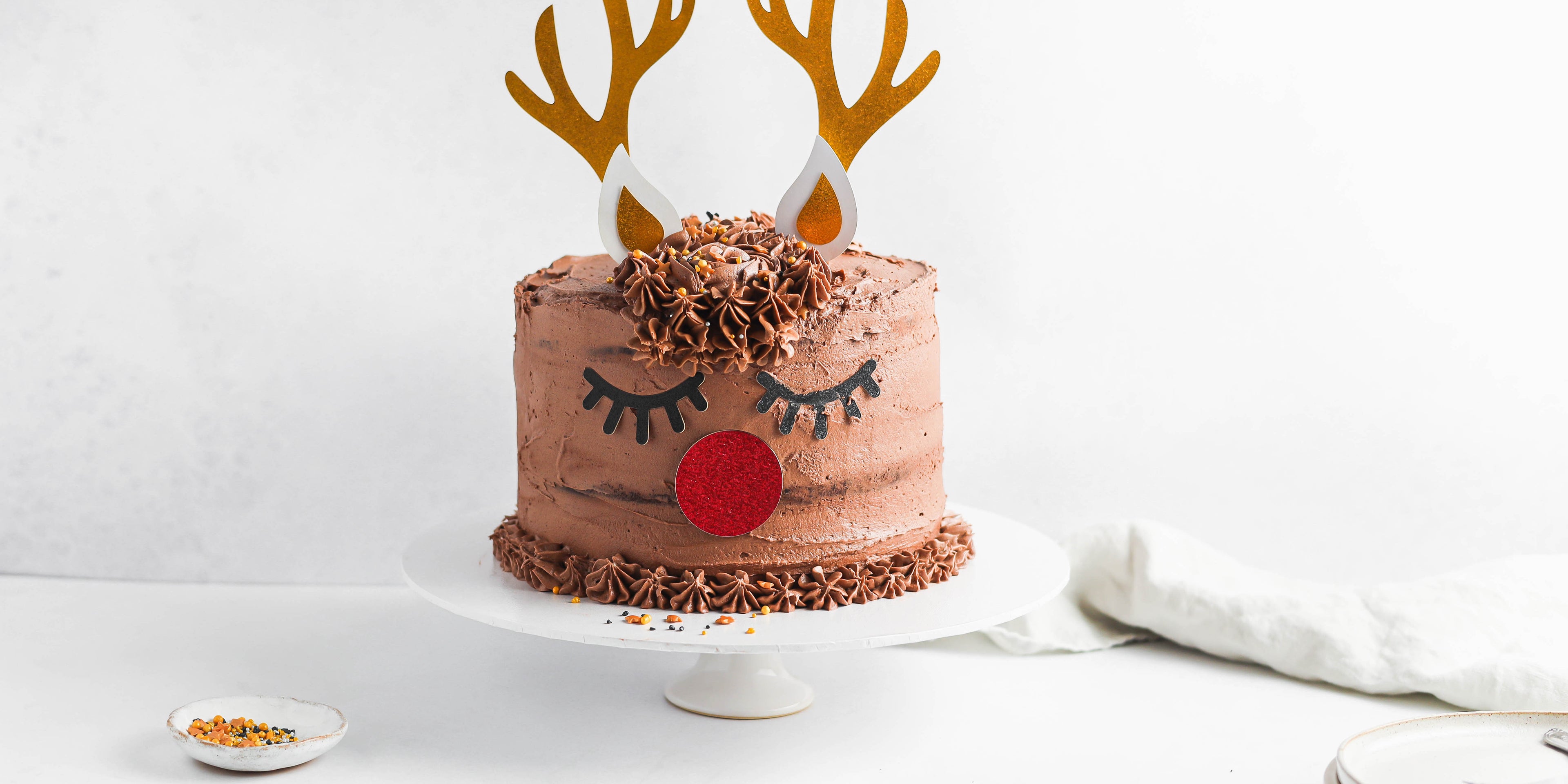 Christmas Reindeer Cake next to a dish of hundreds and thousands on a white cake stand, decorated with golden reindeer antlers