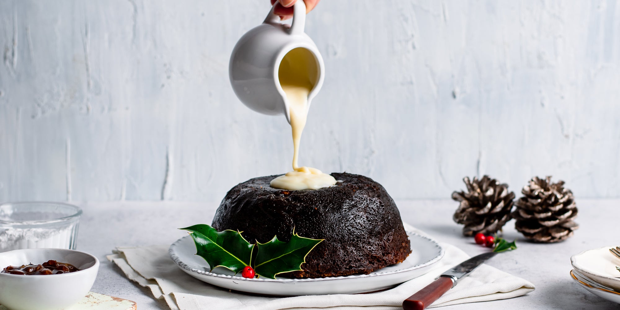 Last Minute Christmas Pudding being poured by a hand holding a jug with cream or brandy butter, next to pine cones and a decorative holly leaf