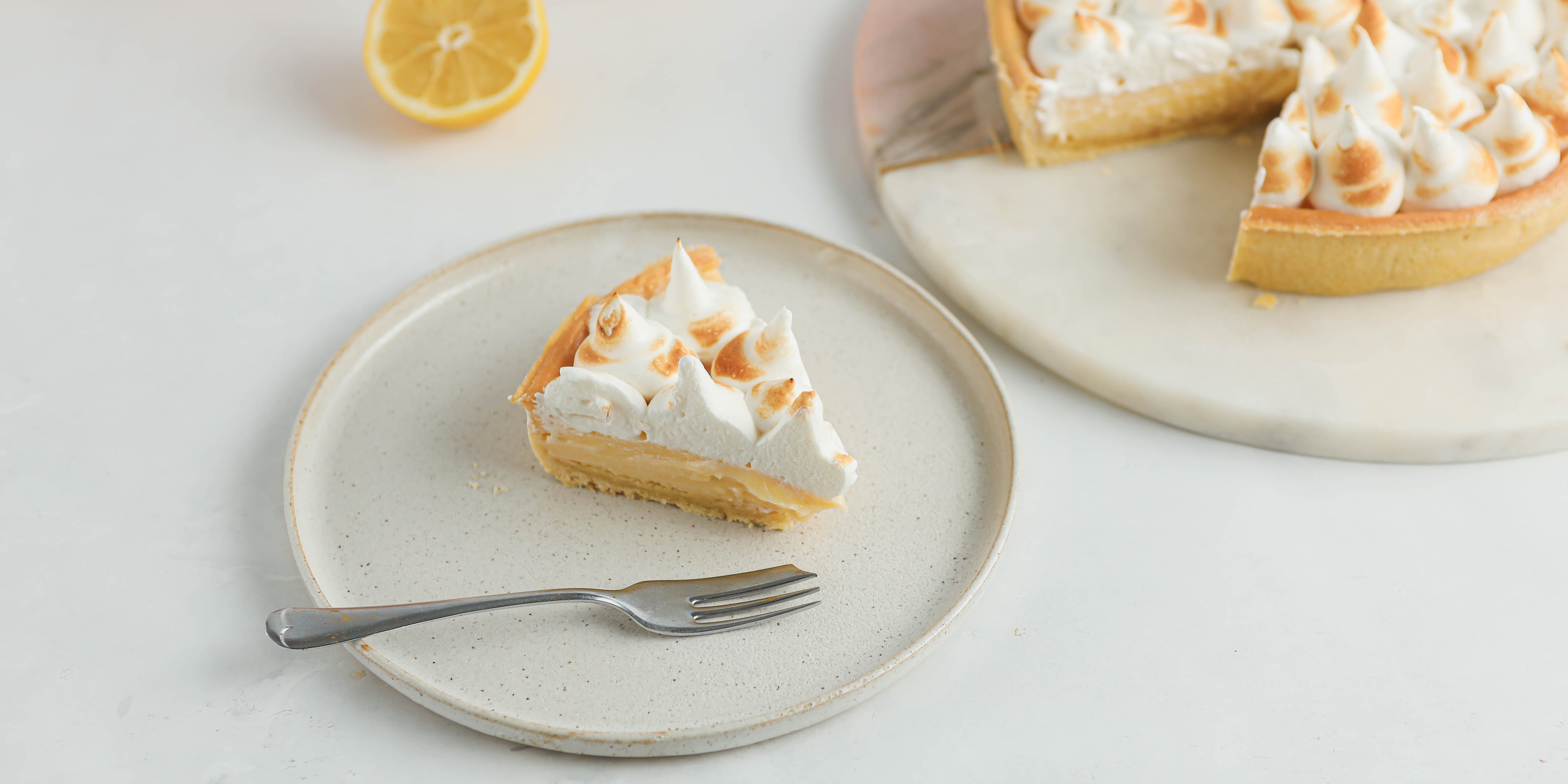 A slice of lemon meringue pie on a white plate with a cake fork. In the background the remaining pie is in shot with a sliced lemon beside it