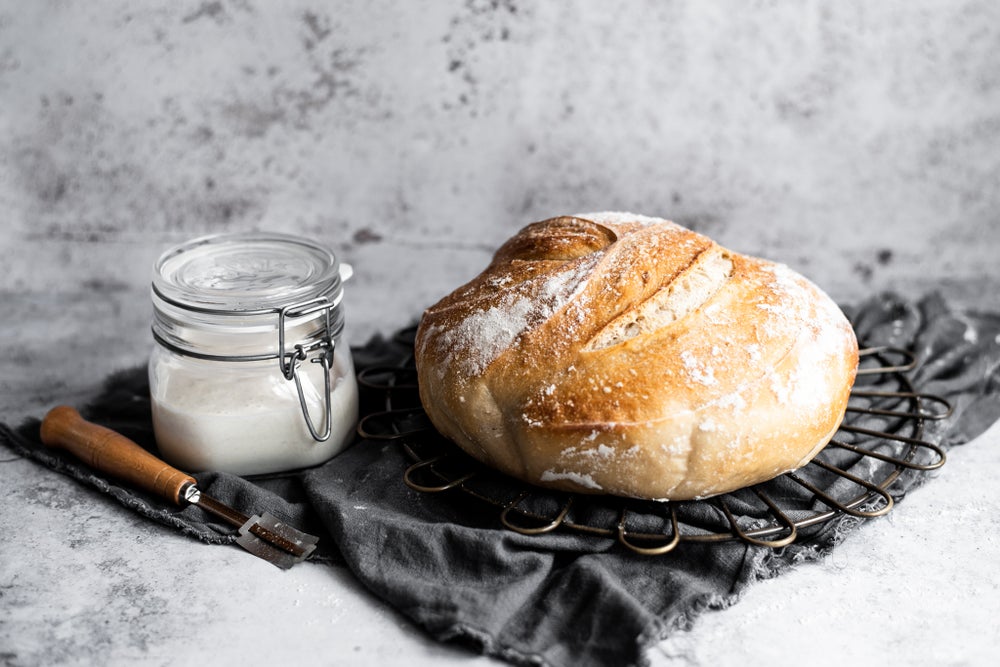 A glass jar of sourdough starter next to a sourdough loaf, placed on some charcoal grey fabric.