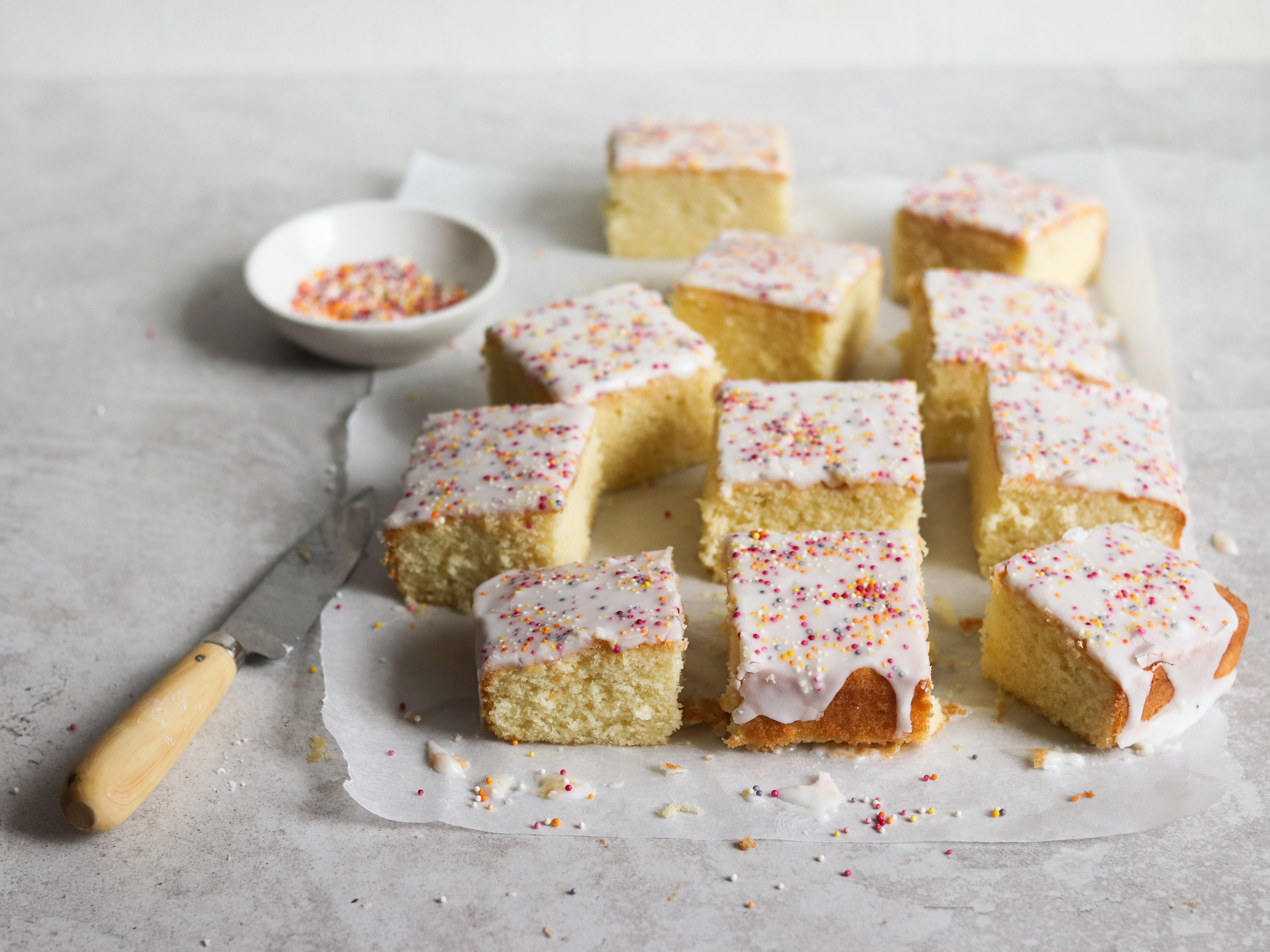 Sprinkle Cake slices with icing and sprinkles dripping down the cake. On a sheet of baking paper next to a knife and a bowl of sprinkles