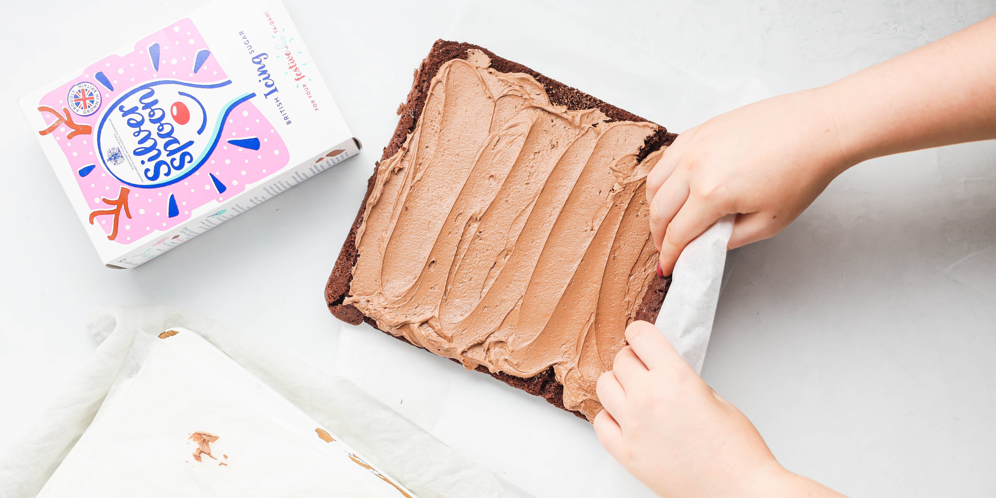Action shot of Chocolate Yule Log being rolled using parchment and hands rolling the cake together. Next to a flat lay box of Silver Spoon icing sugar