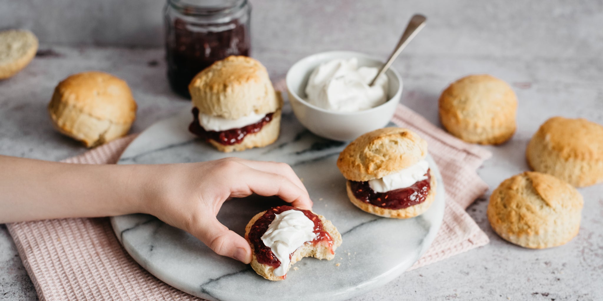 Hand reaching for a Vegan Scone cut in half with jam and plant based whipped cream, next to a bowl of vegan cream and jam