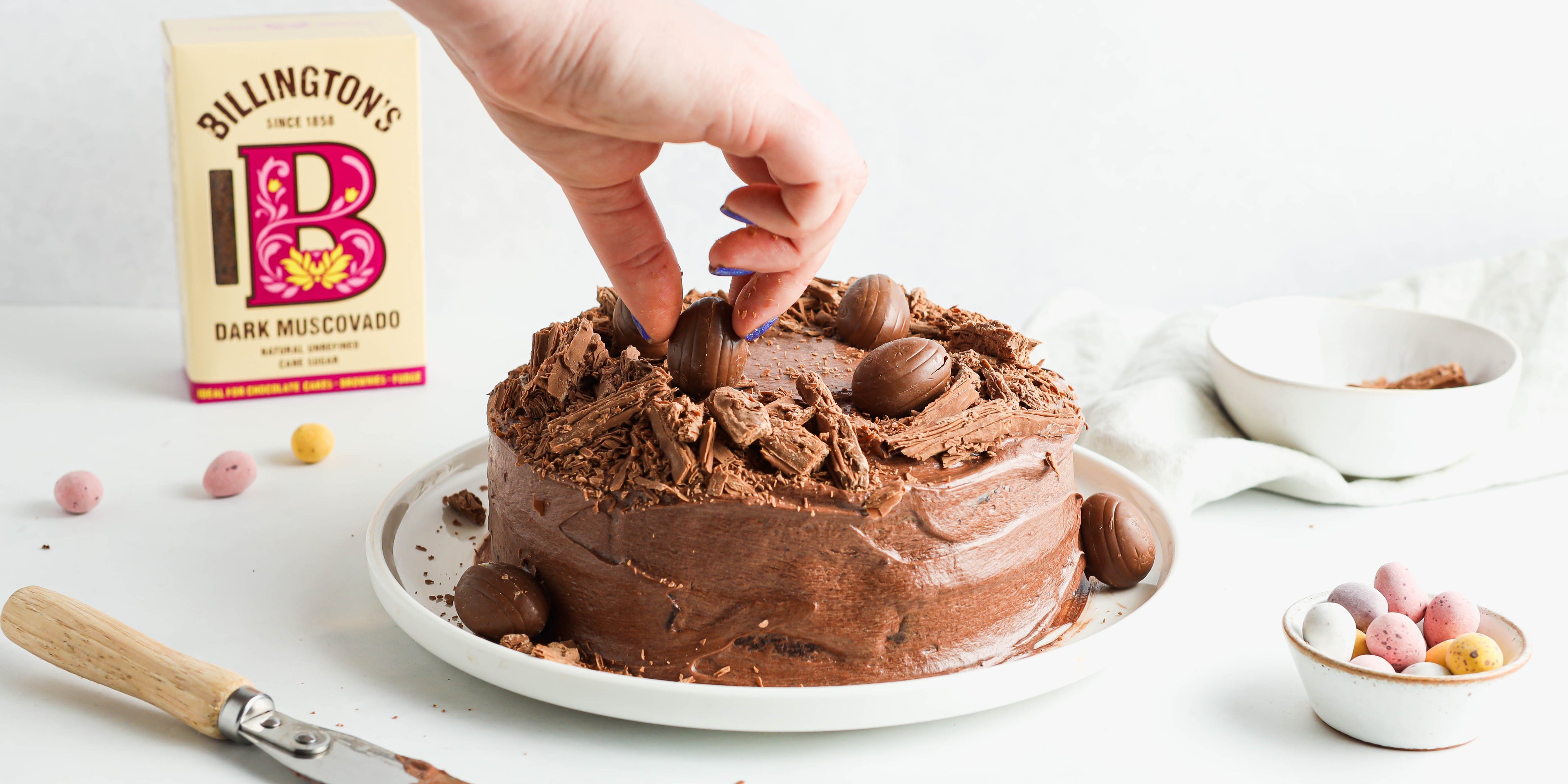 Chocolate Nest Cake with a hand reaching to decorate the top with chocolate eggs, with a box of Billington's Dark Muscovado Sugar in the background