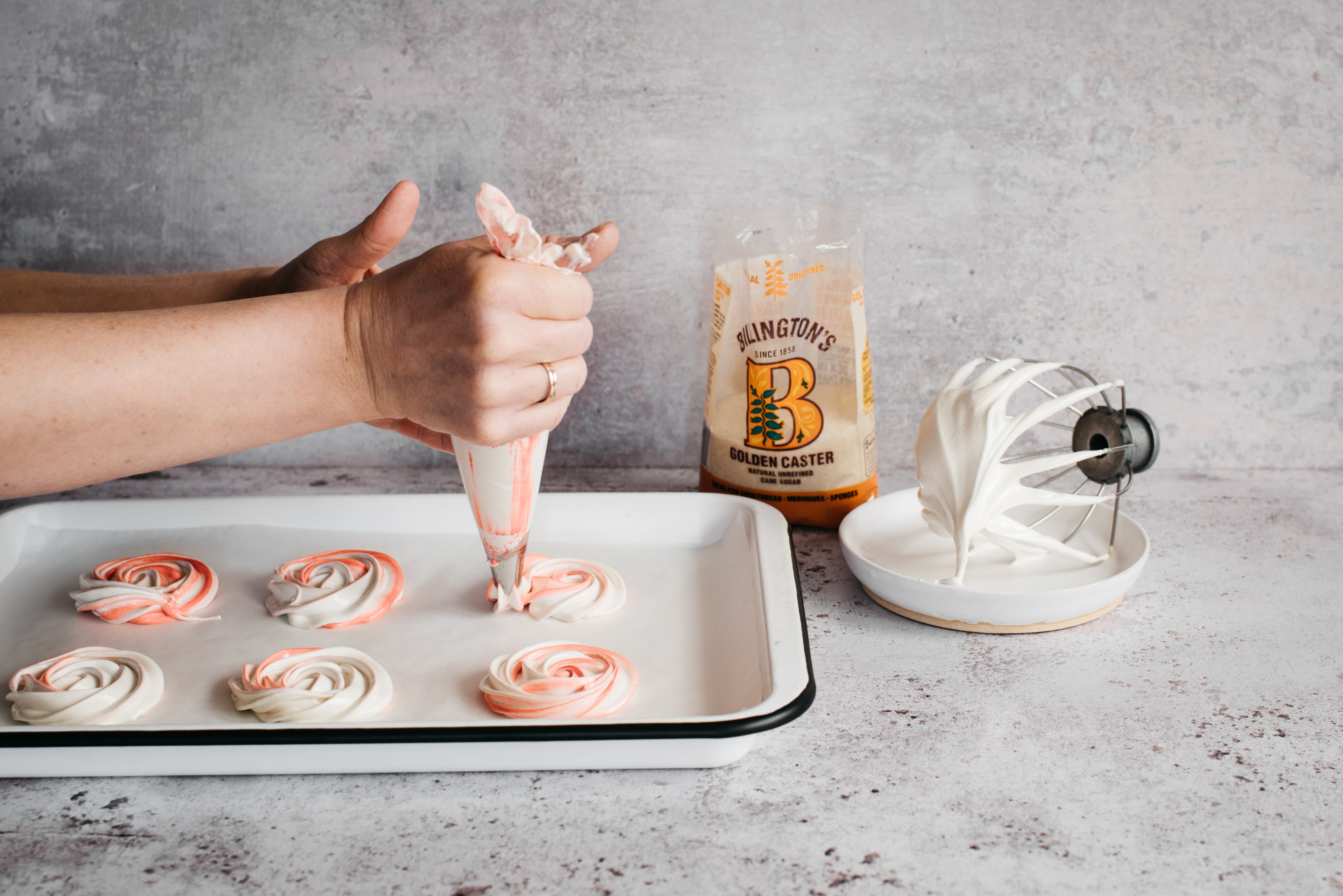 Hand holding a piping bag, piping Vegan Meringue on a baking tray next to a bag of Billington's Golden Caster Sugar