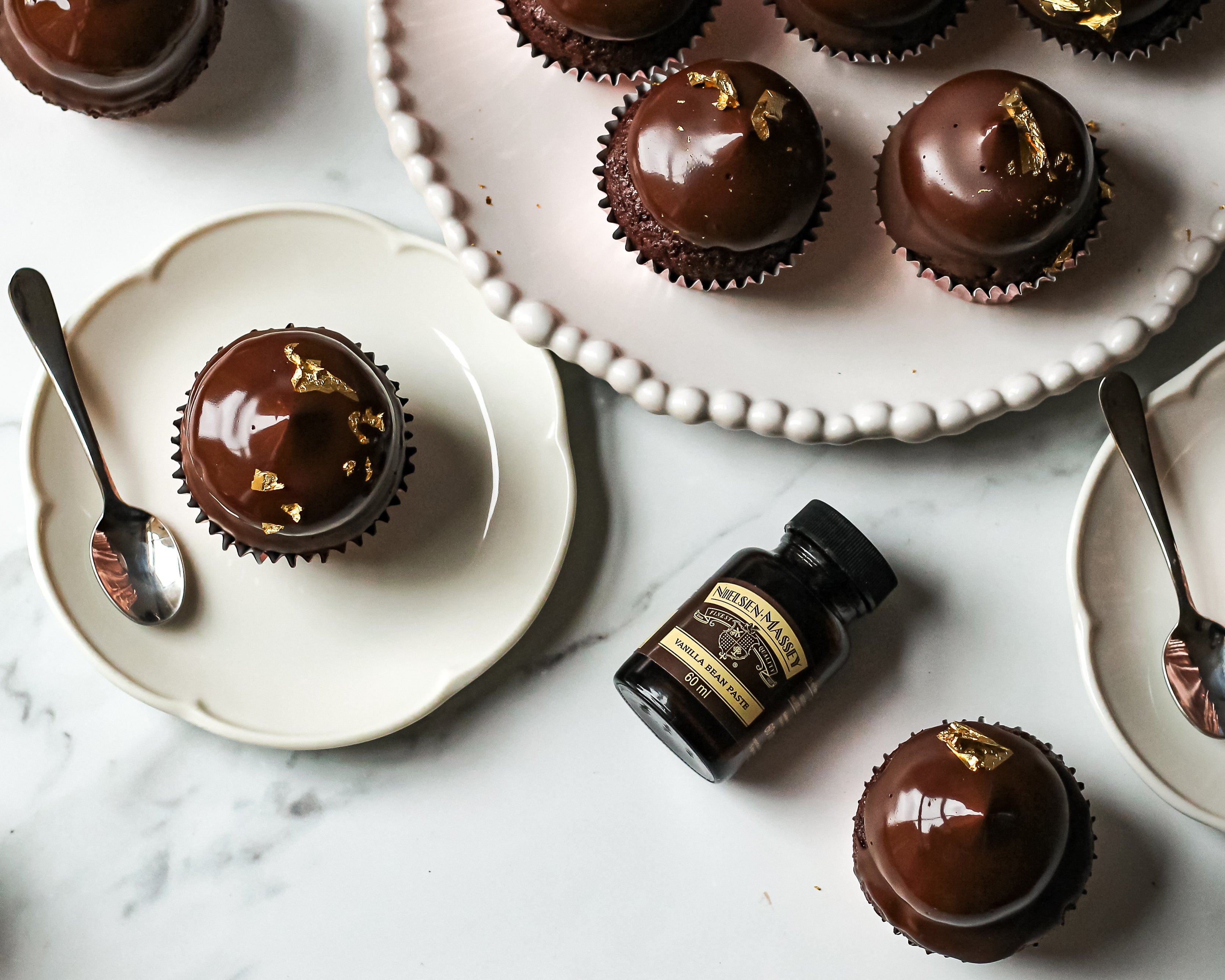 Top view of Hi-Hat Cupcakes on plates ready to serve, next to a flat lay bottle of Nielsen-Massey vanilla extract