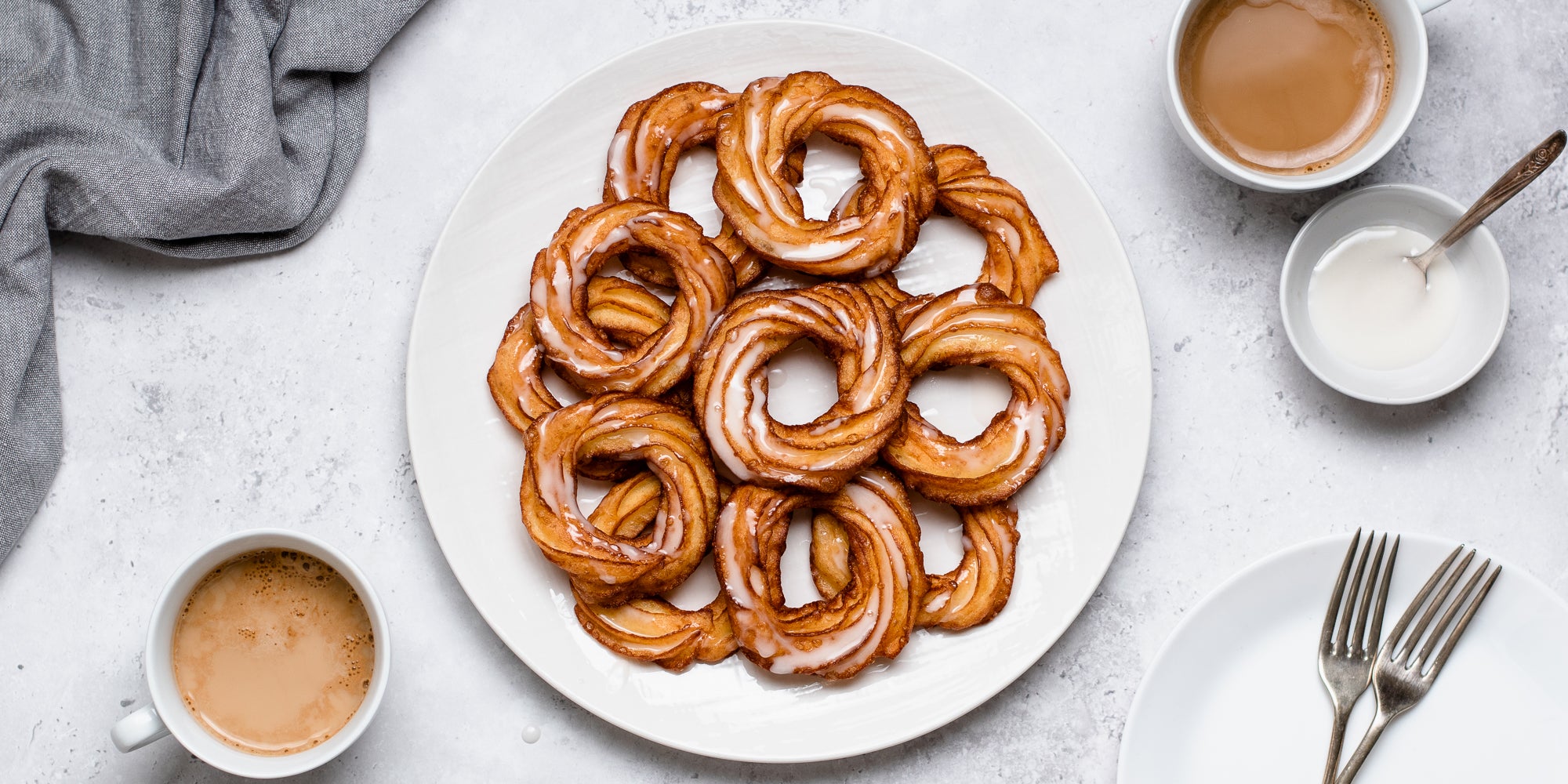 Top view of Apple Cider Crullers on a plate. Next to cups of coffee, and a bowl of icing ready to decorate