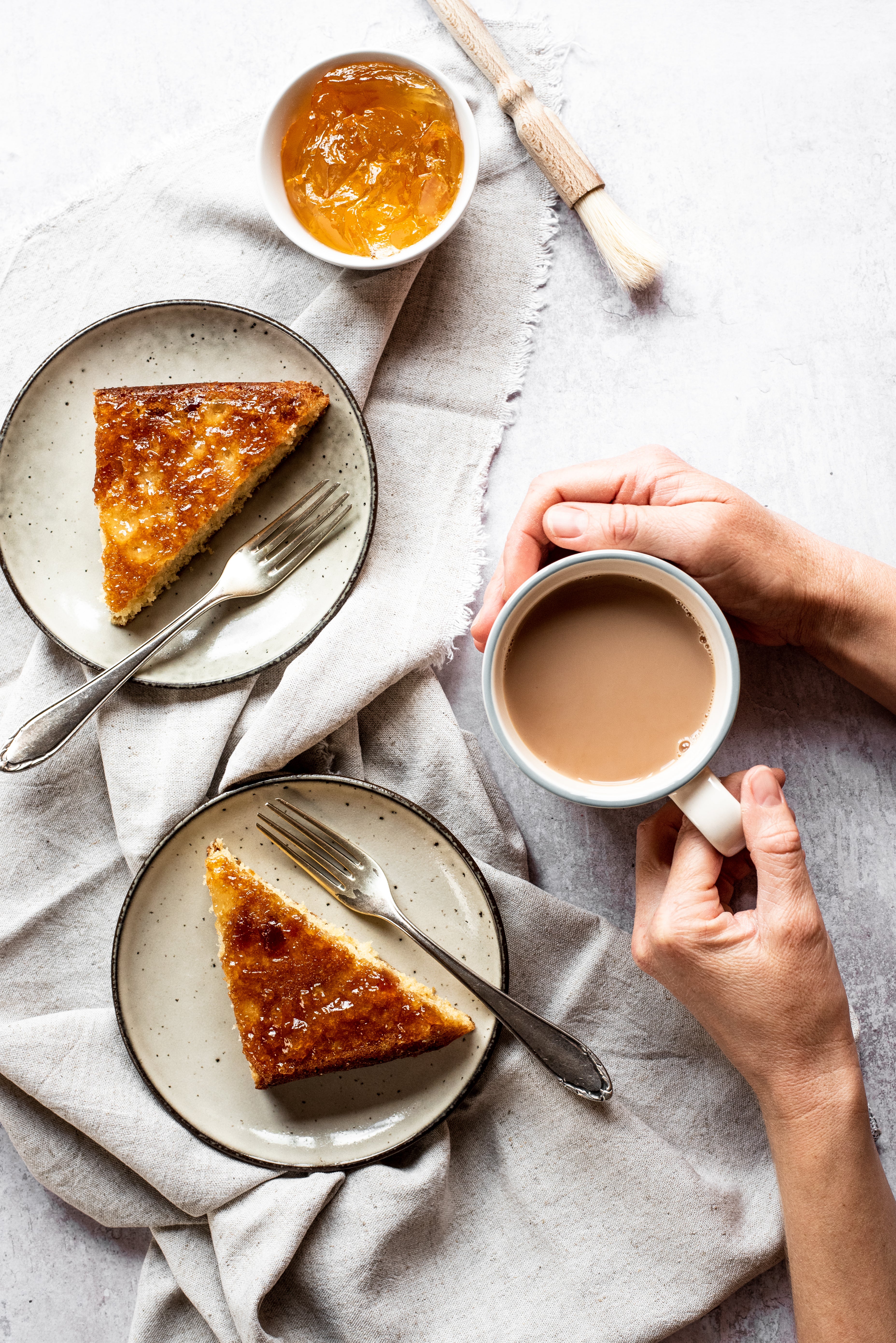Top view of Marmalade Traybake, served on plates with forks. Plates are lay on top of linen cloths, next to hands holding a mug of tea